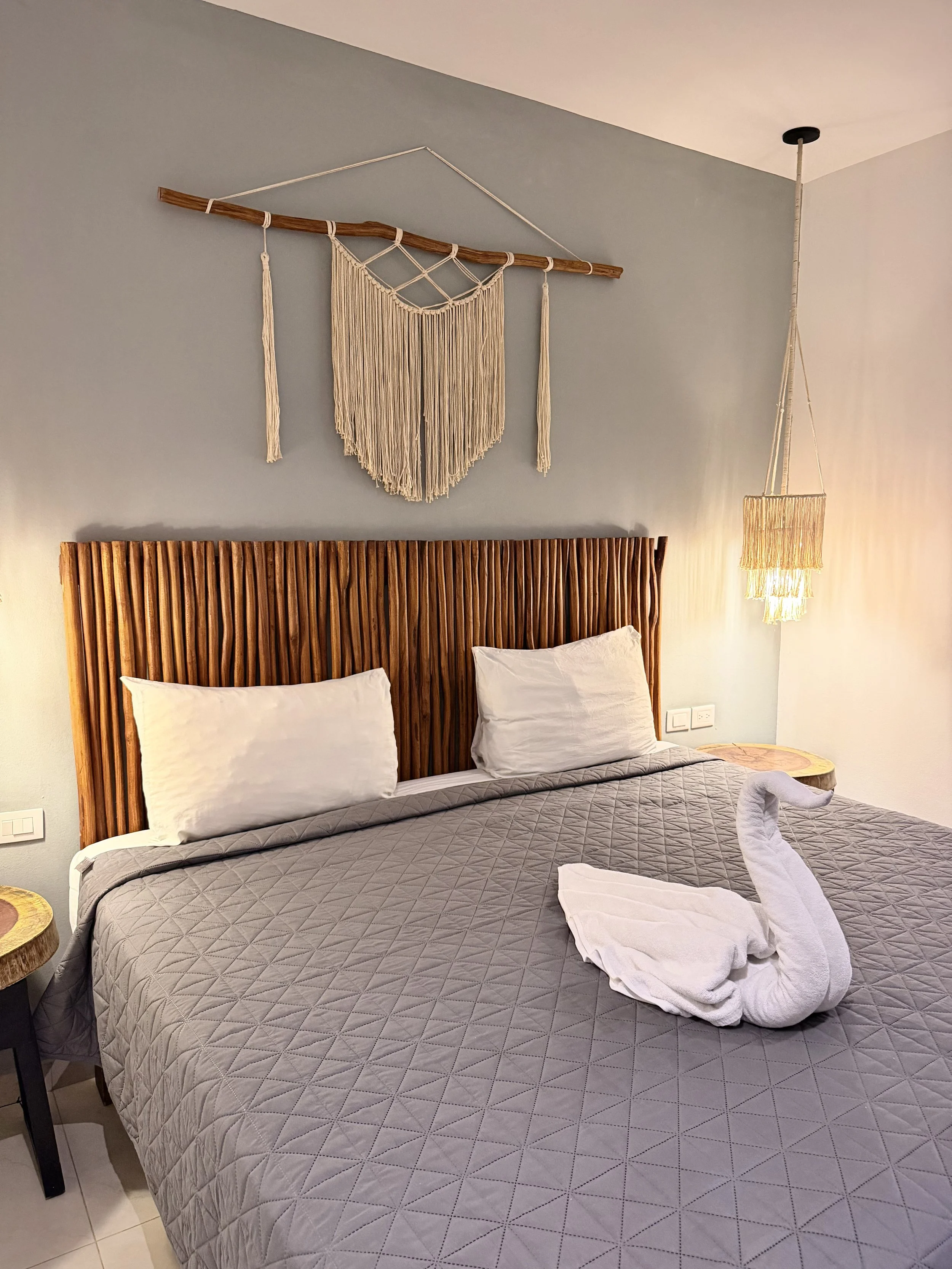 Cama con cabecera de madera, dos almohadas blancas, edredón gris, adorno de toalla en forma de cisne, lámpara de techo de cestería y decoración de macramé en la pared.