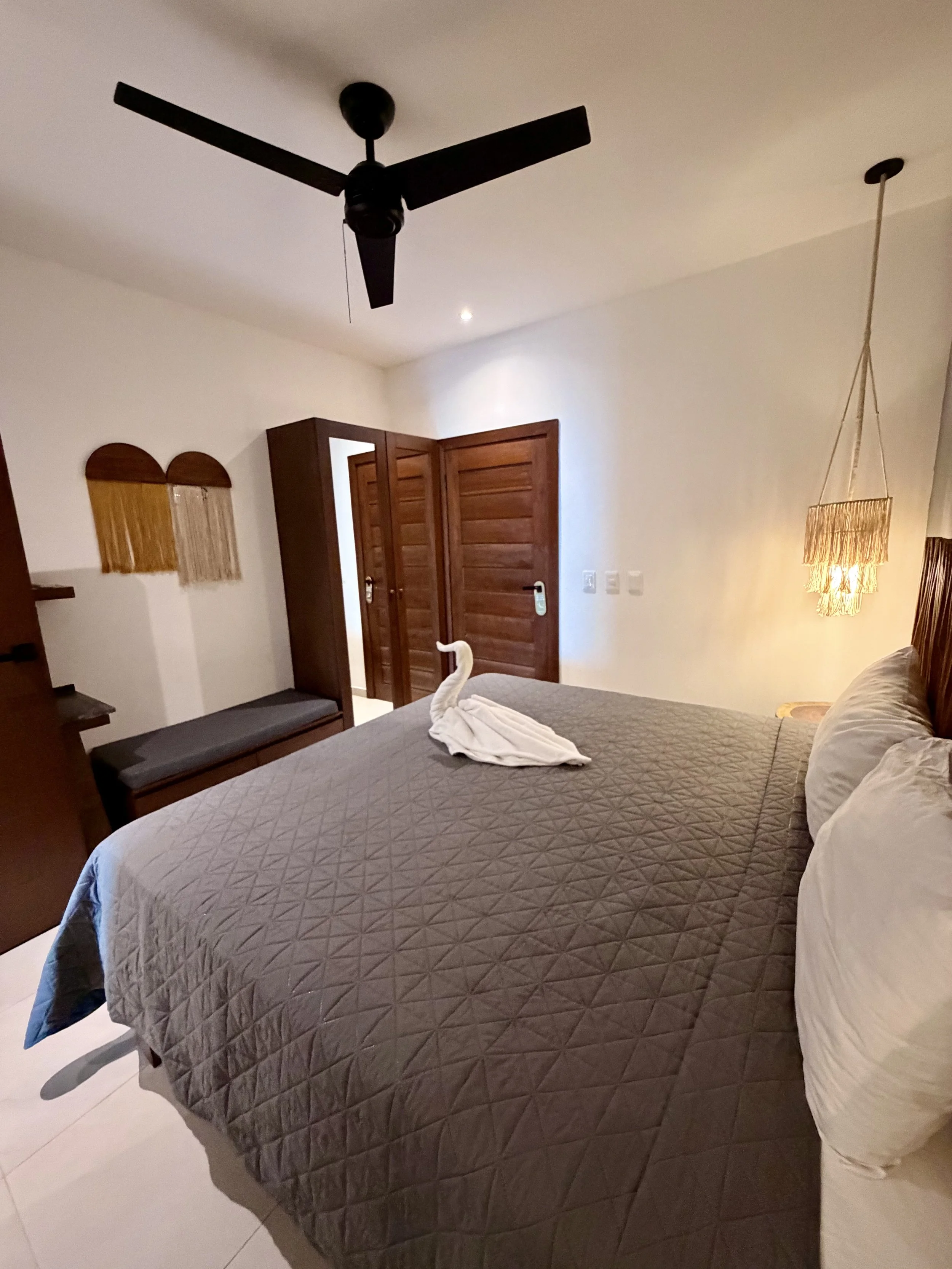 Habitación con cama, toalla doblada en forma de cisne, puerta de madera, lámpara de pared, ventana con cortinas, ventilador de techo y pared blanca.