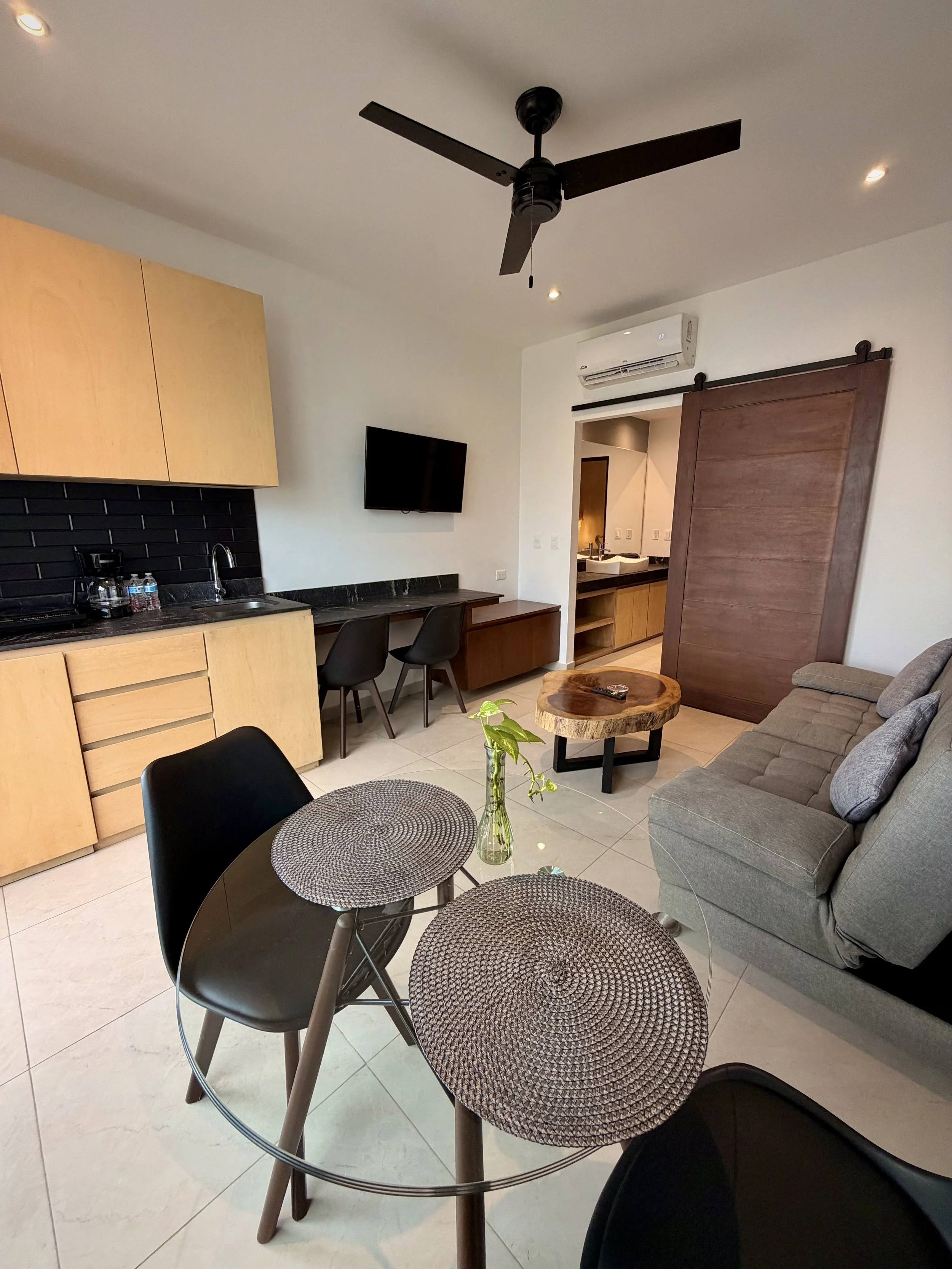 Interior de sala con mesa redonda y sillas, sofá gris, mesa de centro de madera, cocina con fregadero, ventilador de techo, televisor, aire acondicionado y puerta corredera de madera.