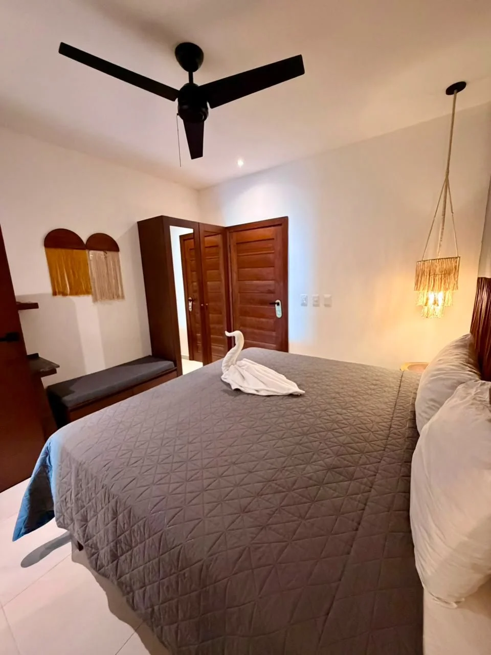 Habitación con cama doble, colcha gris, cortinas de la pared en forma de arco, lámpara de pared, ventilador negro en el techo, puerta de madera abierta, y una toalla doblada en forma de cisne sobre la cama.