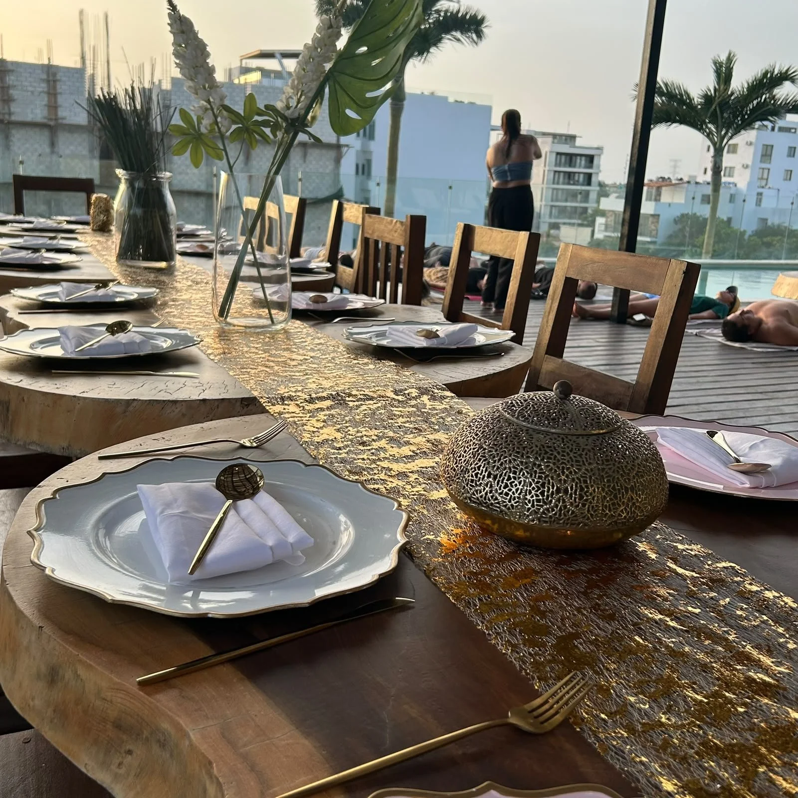 Mesa decorada para una comida o evento con platos blancos, servilletas blancas dobladas, cubiertos dorados y vasos con flores, en un espacio con vista a edificios y palmeras, y personas haciendo yoga en el fondo.