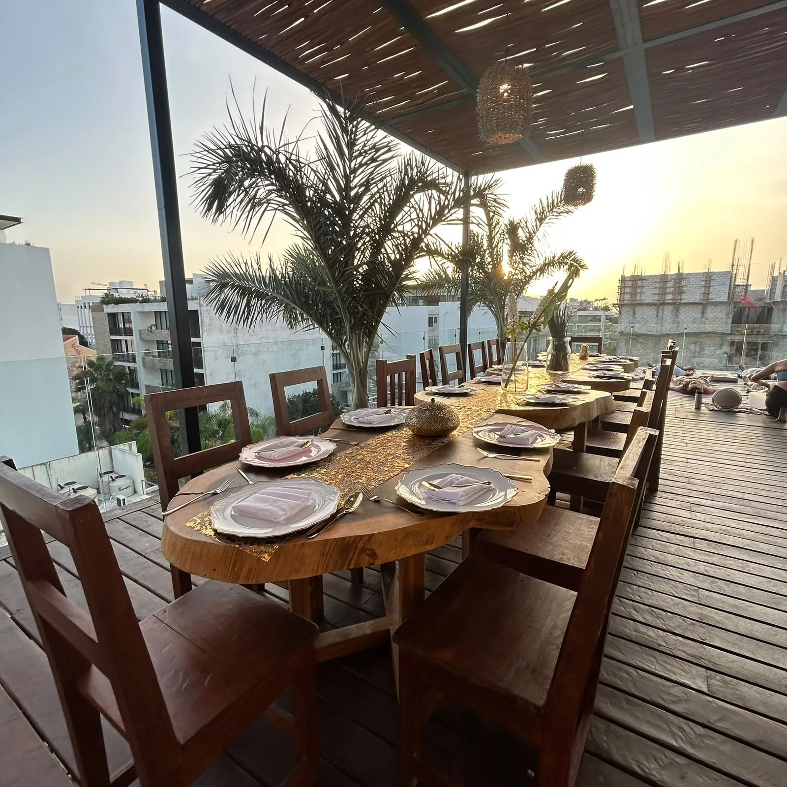 Mesa larga de madera con platos, cubiertos y servilletas, en una terraza con vista al atardecer, plantas y decoración