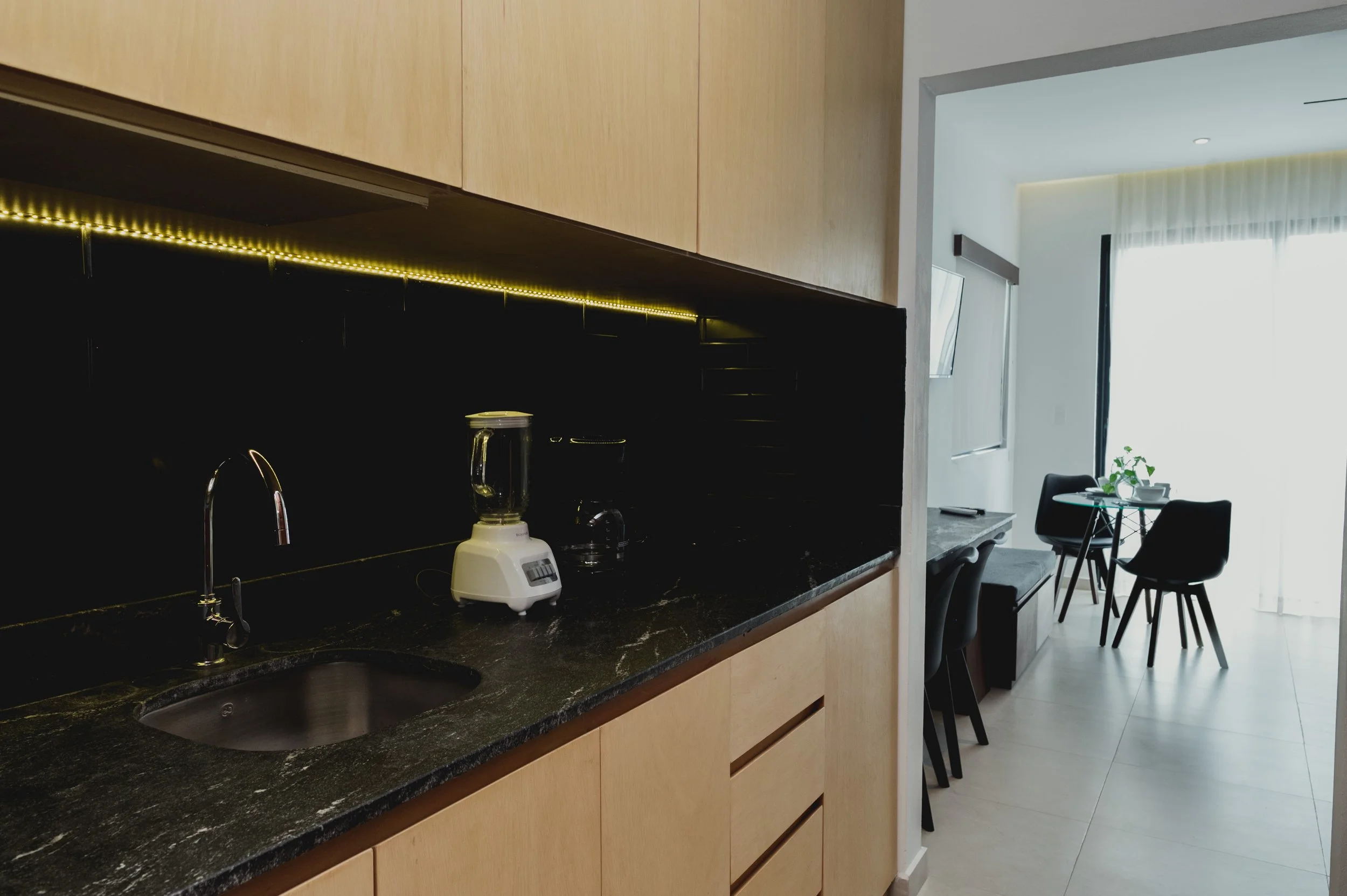 Vista de una cocina moderna con encimera de granito negro, armarios de madera clara y un fondo oscuro con iluminación LED. Al fondo, se observa un área para comer con sillas negras y una mesa redonda, cerca de una ventana con cortinas blancas.