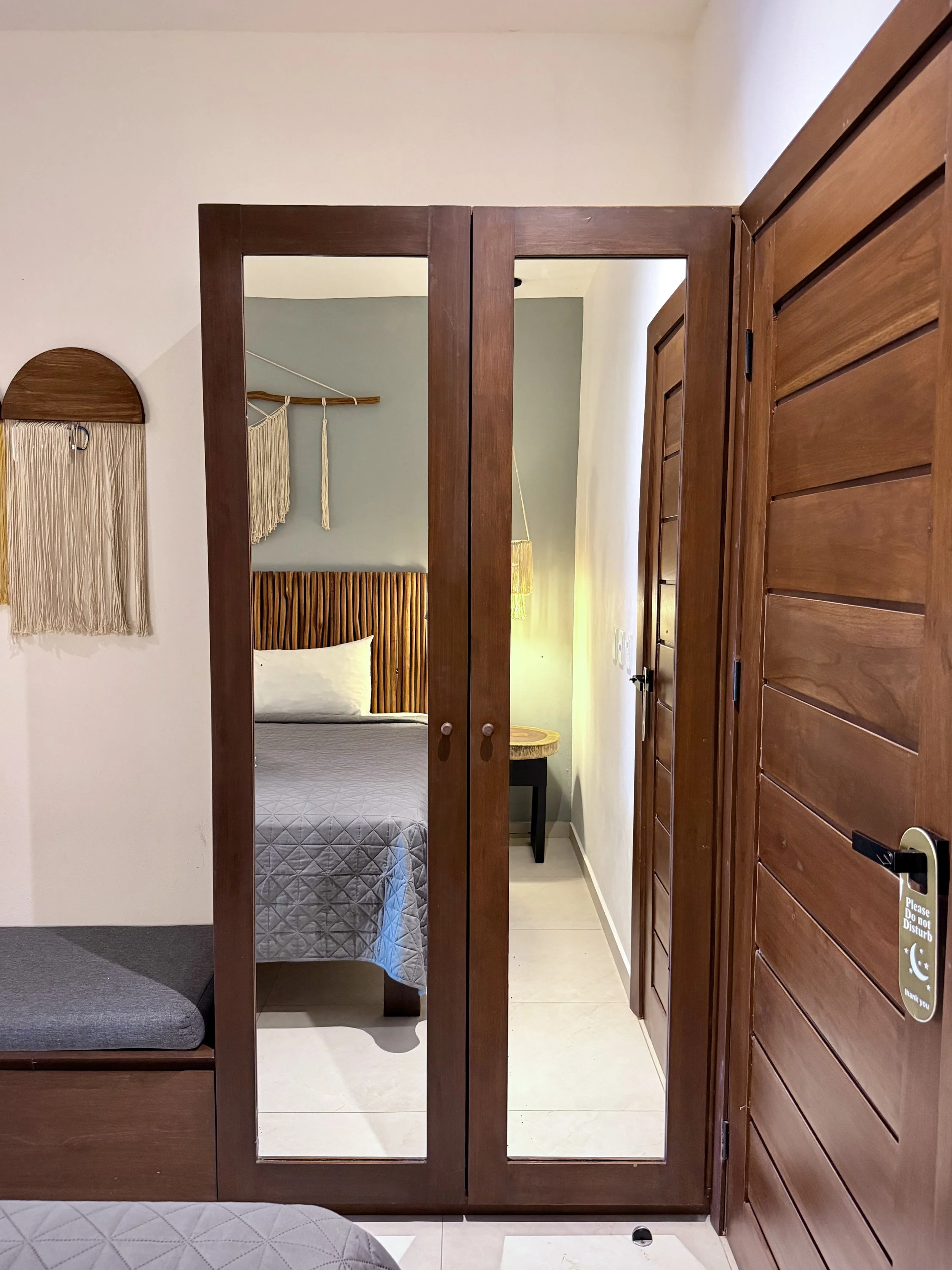 Puerta de closet de madera que refleja la habitación, mostrando una cama con cabecera de madera y una lámpara de pie al fondo, con paredes de color verde.