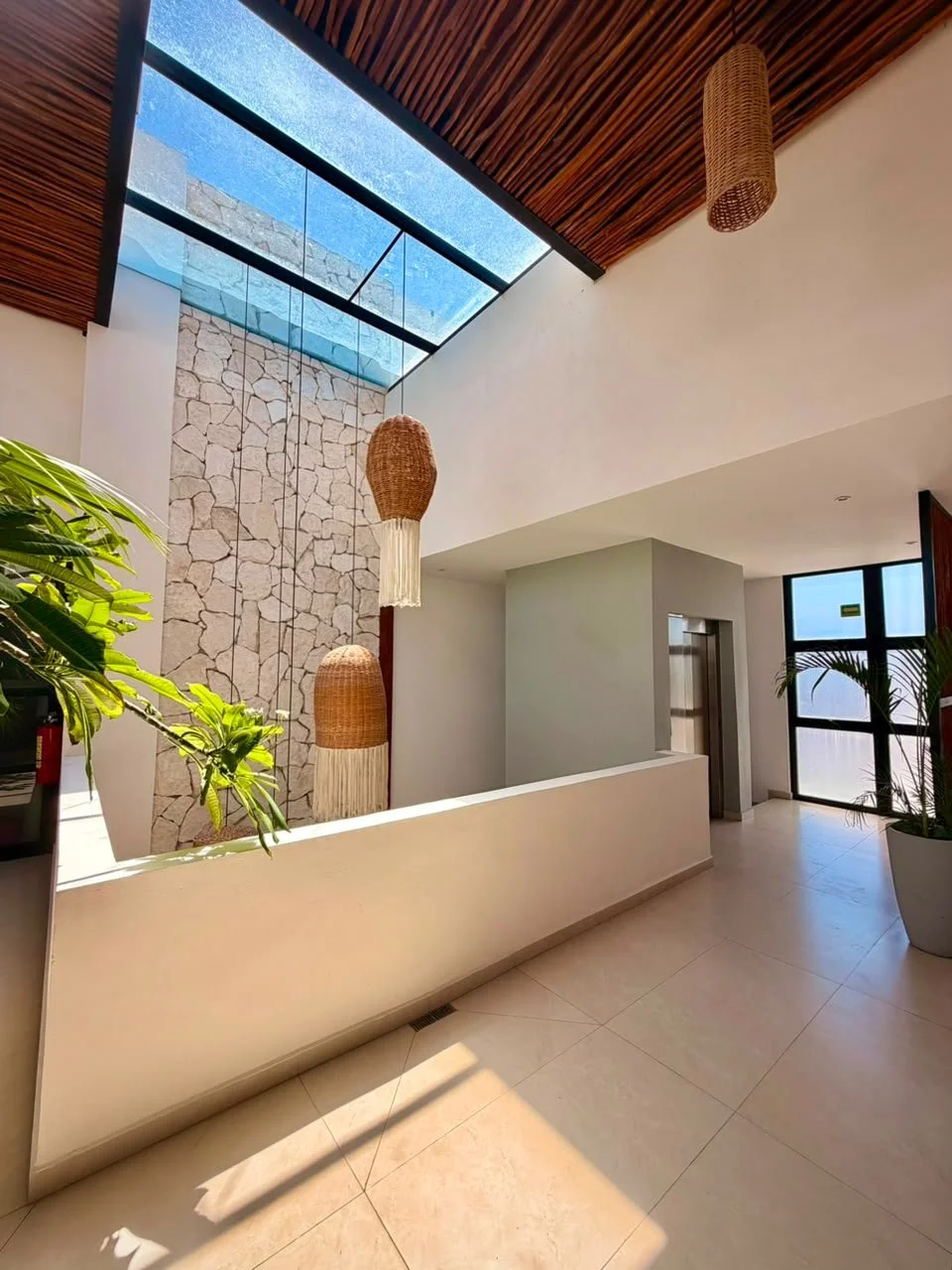 Interior moderna con muro de piedra, techo de madera, lámparas colgantes de mimbre, paredes blancas, plantas verdes y ventanas grandes con vista al exterior.