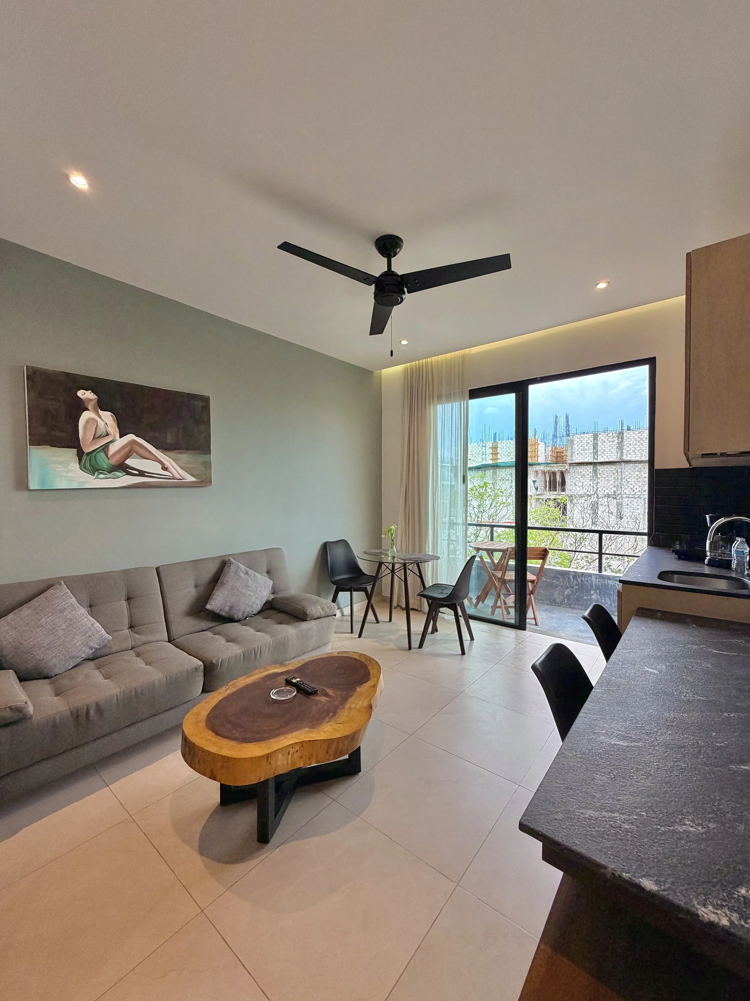 Salón con sofá gris, mesa de madera en forma de río, mesa de comedor redonda con sillas negras, ventana con vista a edificios en construcción, pintura en la pared y un ventilador negro en el techo.