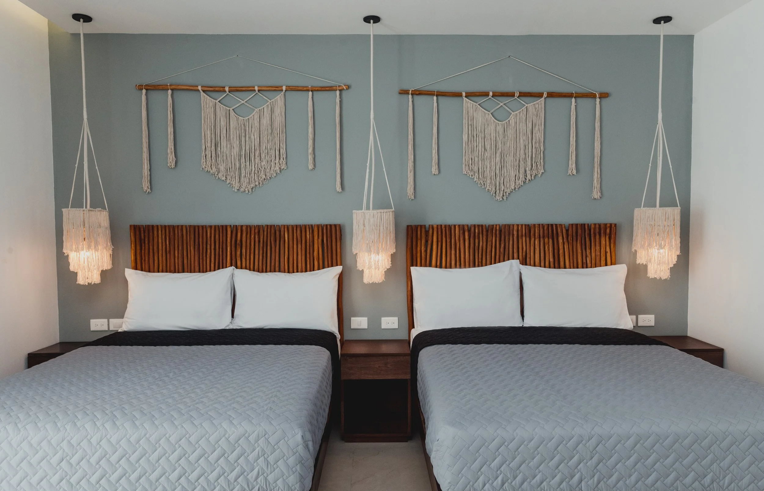 Habitación con dos camas, cabeceras de madera, lámparas colgantes, y decoraciones de macramé en la pared.