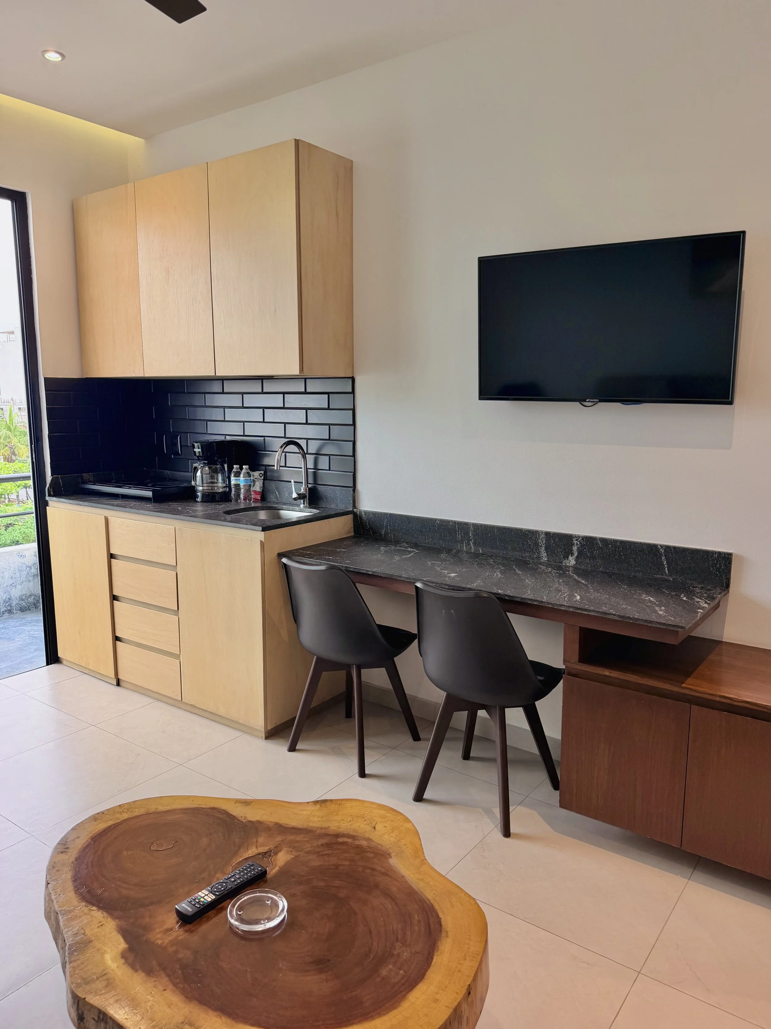 sala y cocina con muebles de madera clara, una mesa con top gris oscuro, sillones negros, televisor en la pared, y un cerdo de madera en primer plano con control remoto, cenizas y vaso en la mesa.