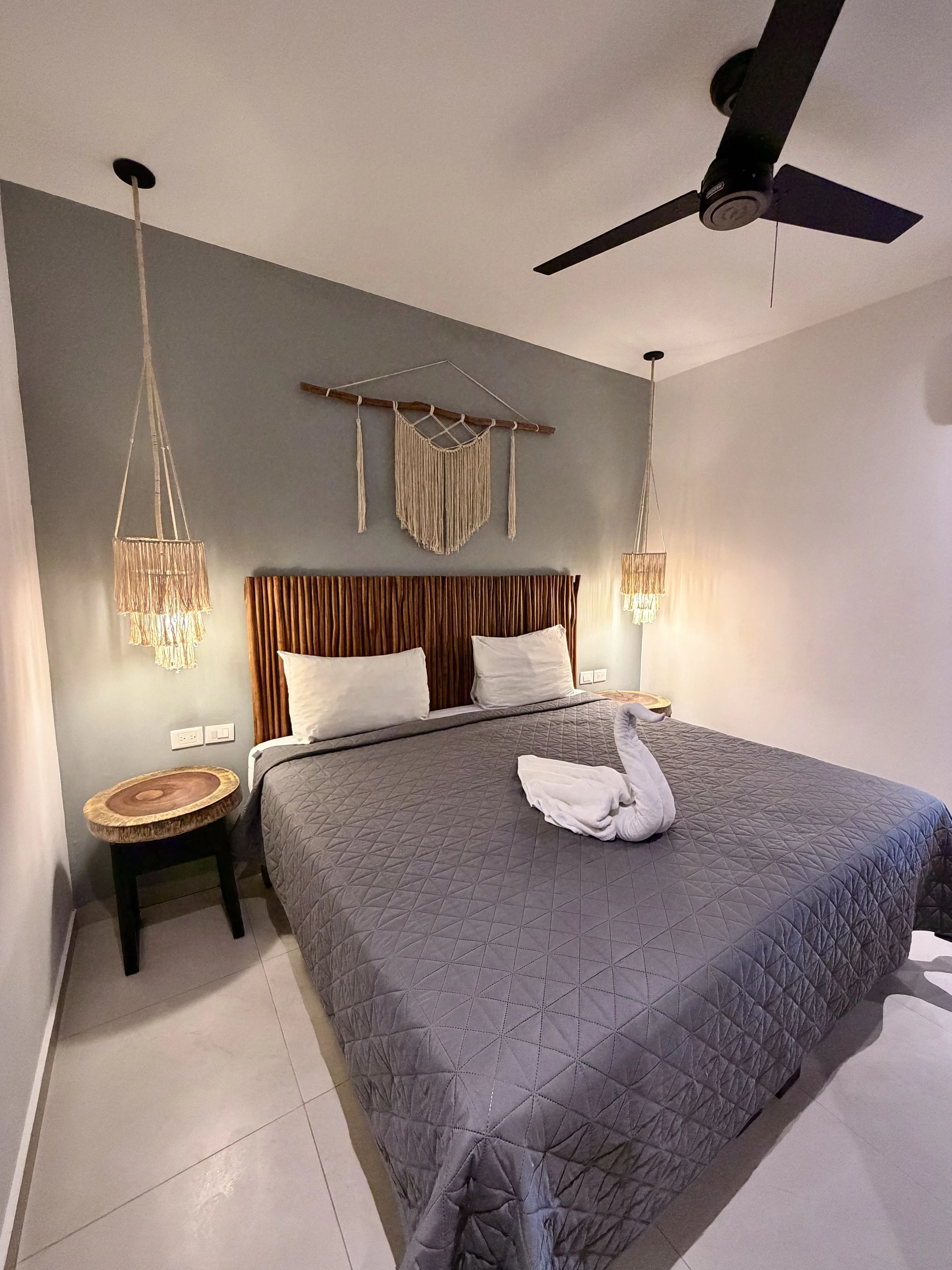 Habitación con cama con cabecera de madera, dos almohadas, lámparas de fibras colgando, pared azul y decoraciones de macramé en la pared, teto con ventilador y paredes blancas, suelo de baldosas claras.
