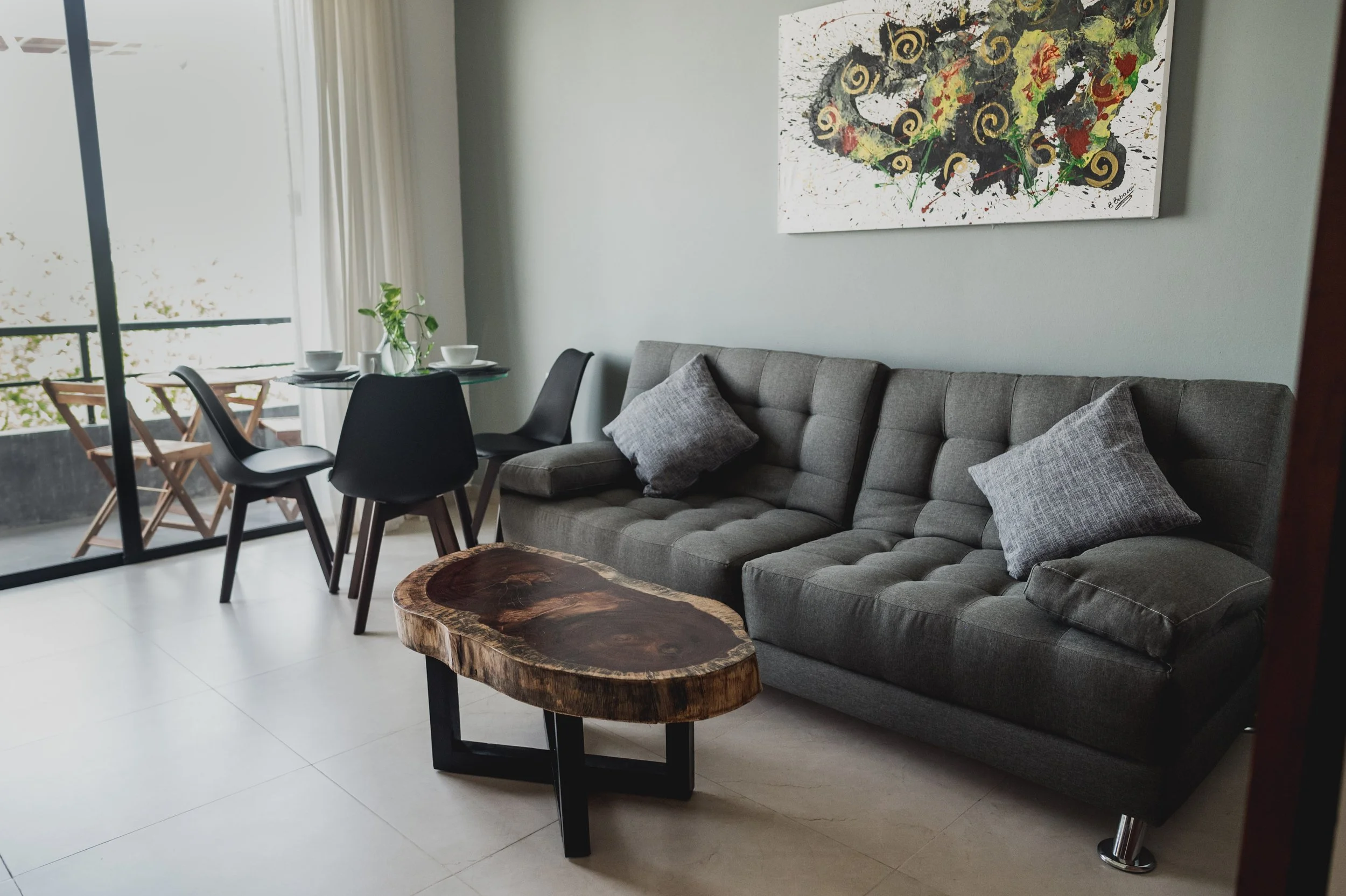 Sala de estar con sofá cama, mesa de madera natural y cojines grises, con cuadro abstracto en la pared y ventana con cortinas blancas, área de comedor con mesa y sillas y balcón con vista a áreas verdes y a la quinta avenida.