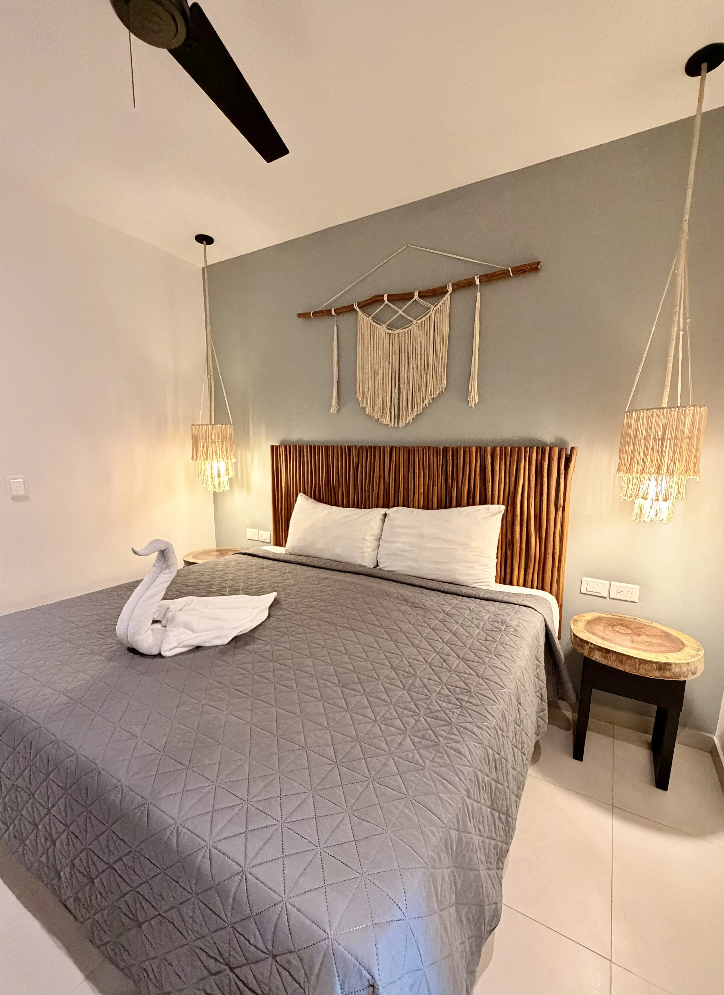 Cama con cabecera de madera, decoración con un tapiz de macramé en la pared, cortinas de lámparas de fibra y toalla en forma de cisne sobre la cama.