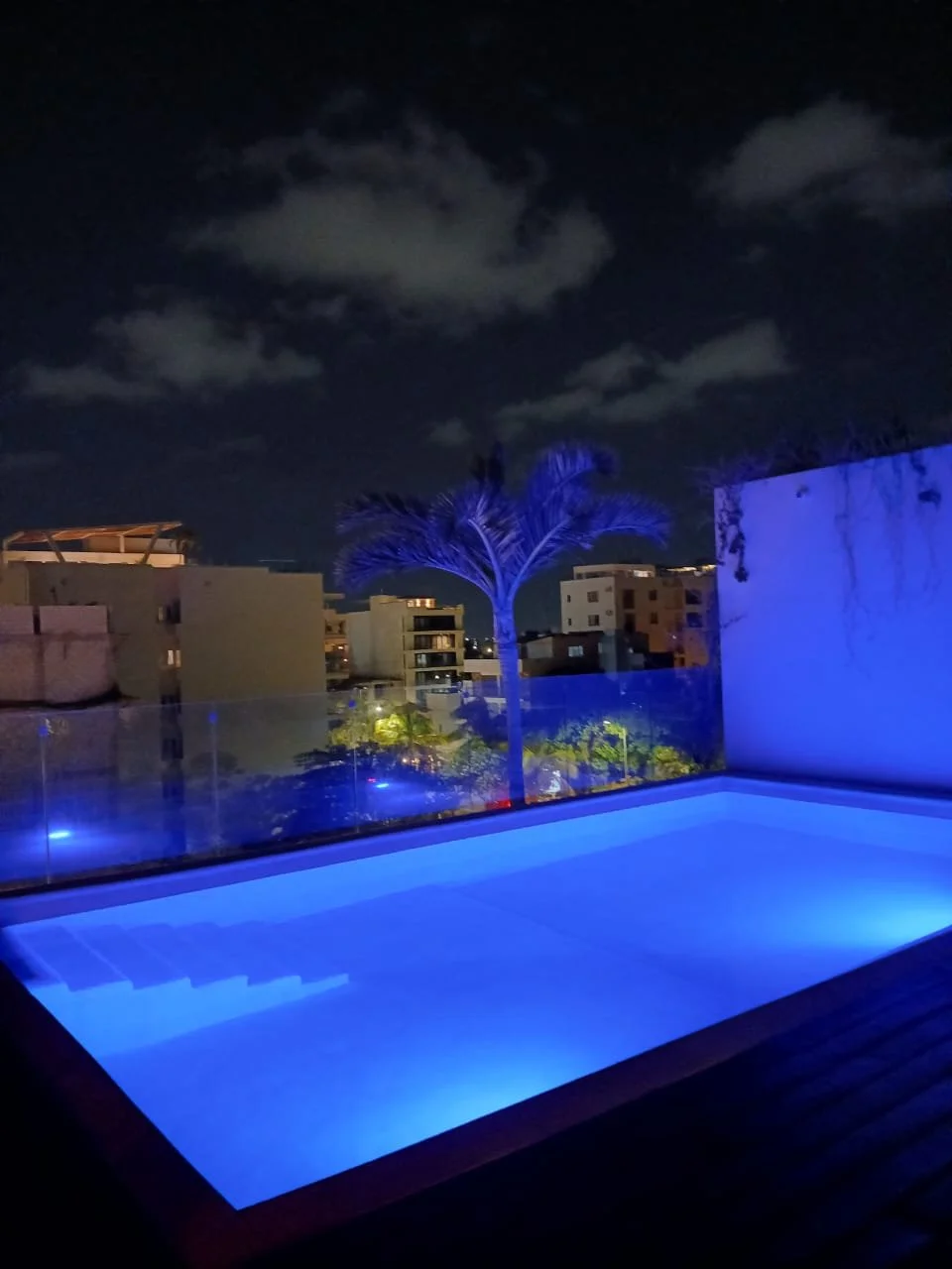 Piscina iluminada con luz azul en una terraza por la noche, con árboles y edificios de fondo bajo un cielo nublado.