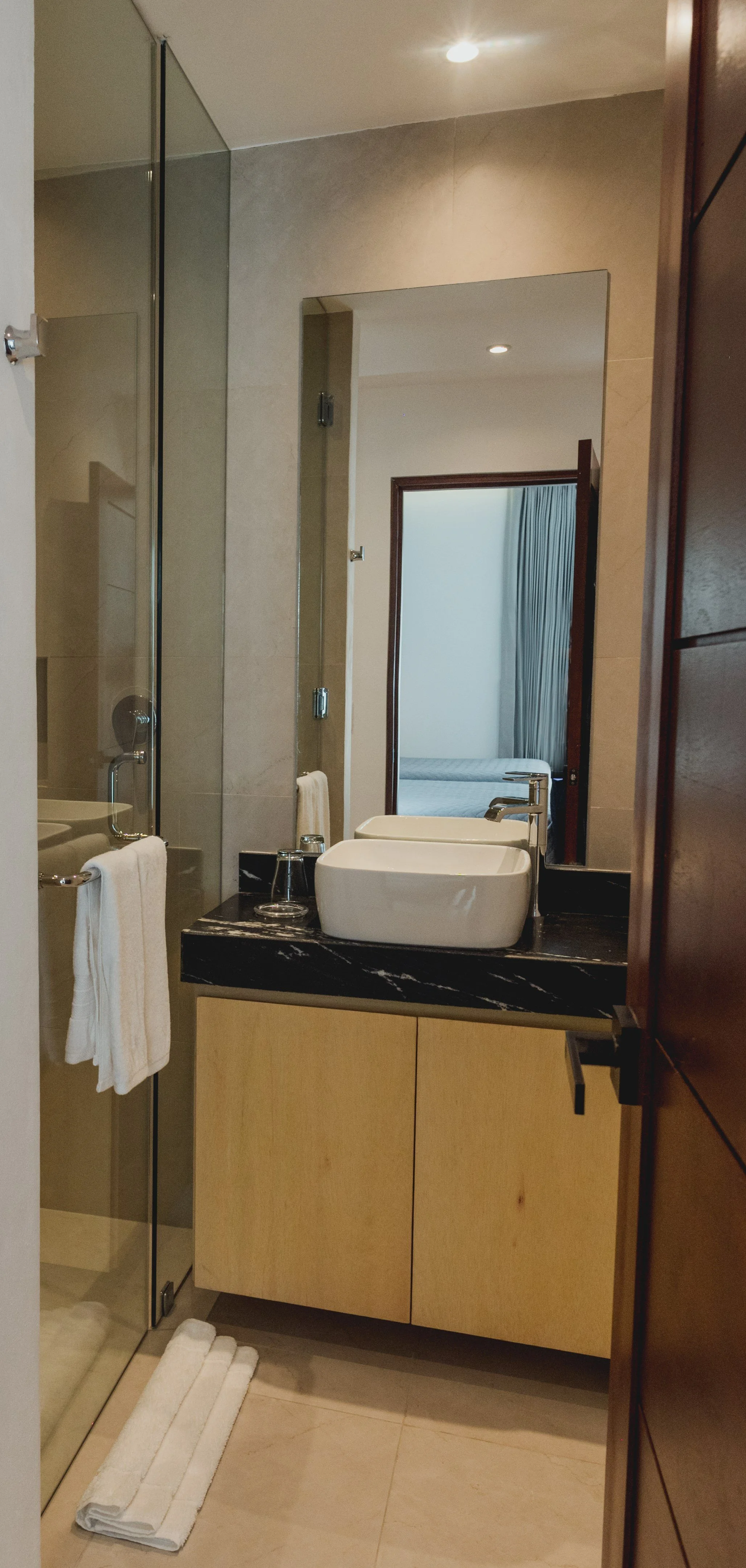Vista de un baño con lavabo, espejo, ducha con vidrio y cuarto habitación visible a través de la puerta abierta.