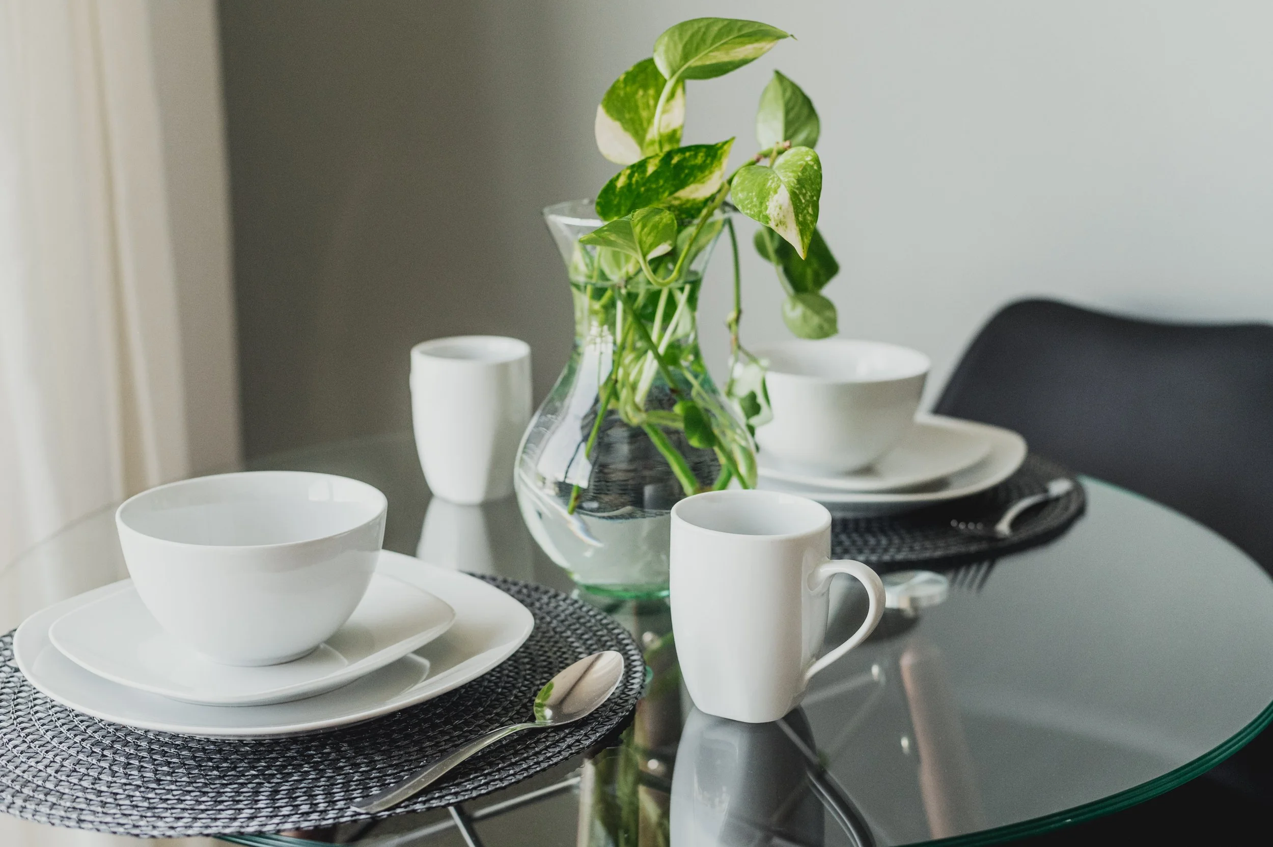 Mesa de comedor decorada con platos blancos, tazas, cucharas, un florero con plantas verdes y paw en un entorno interior con paredes grises.