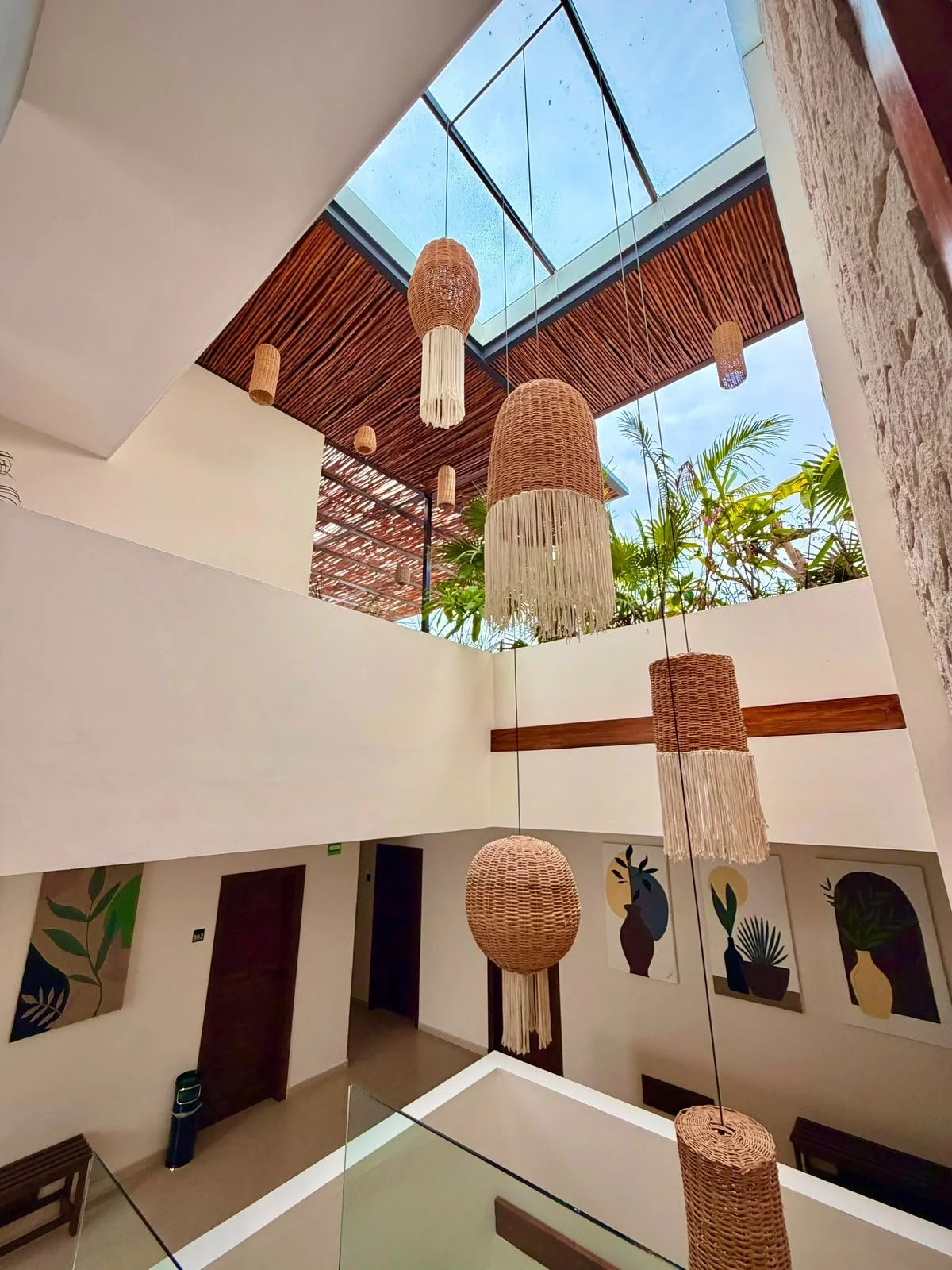 Interior moderno con lámparas de fibra natural colgantes, paredes blancas, arte de plantas en las paredes, y un techo de vidrio con vista al cielo y plantas verdes.