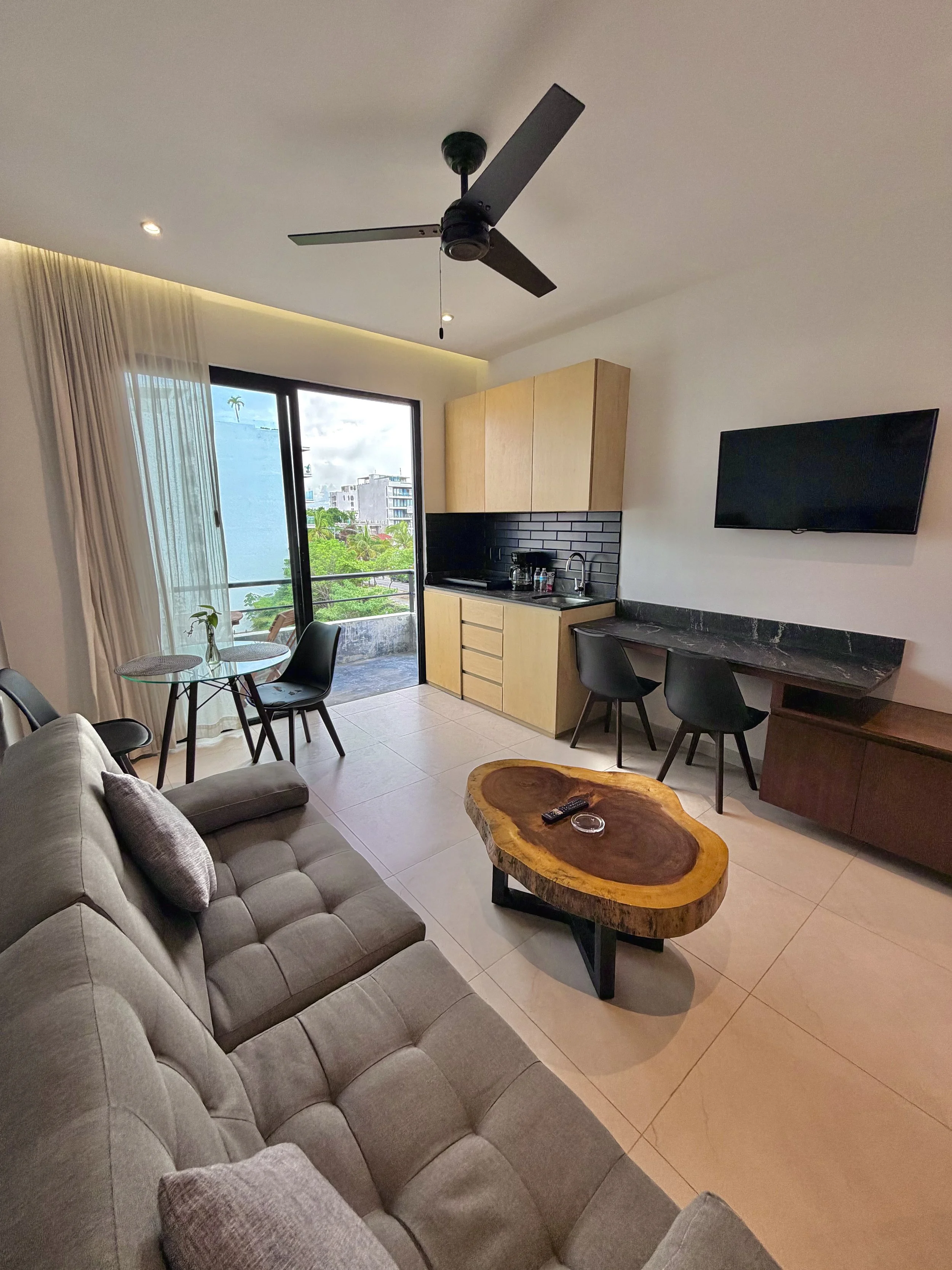 Sala de estar con sofá gris, mesa de madera y varias sillas negras, cocina con gabinetes de madera y pared de ladrillo negro, televisión en la pared, puerta de cristal que da a un balcón con vista a edificios y árboles, cortinas claras.
