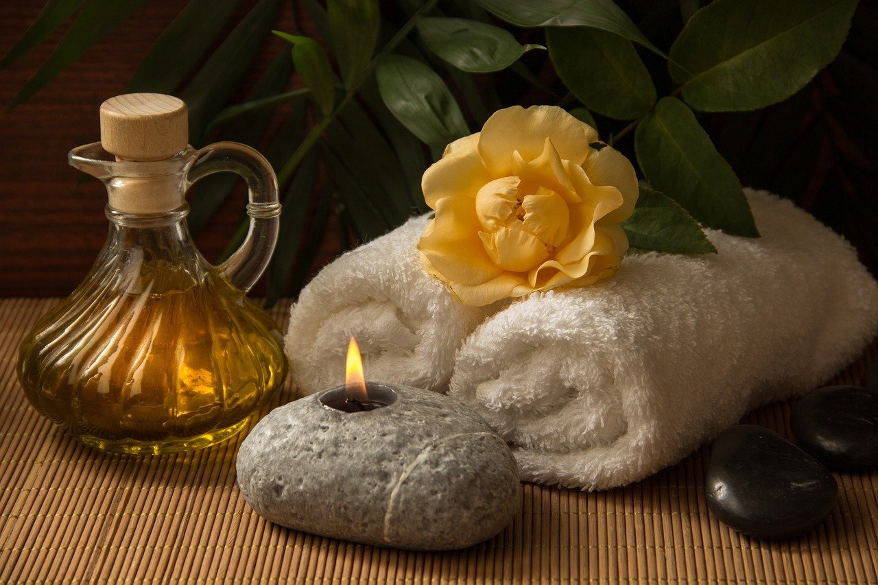Decoración de spa con toallas blancas enrolladas, una flor amarilla, piedras negras, una vela encendida y una botella de aceite con tapón de madera sobre una estera de bambú.