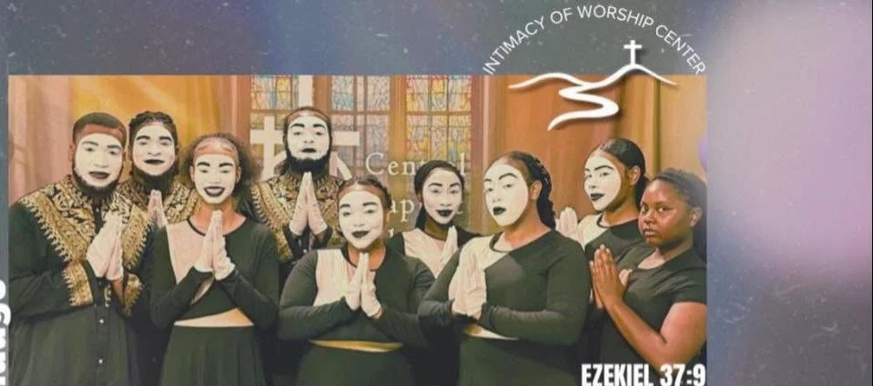 Group of nine people dressed in black and gold religious attire standing inside a church with stained glass windows, some with painted faces, praying or standing in prayer posture, near a sign that reads 'Community of Worship Center' with a cross and mountain illustration, and a Bible verse Ezekiel 37:9.
