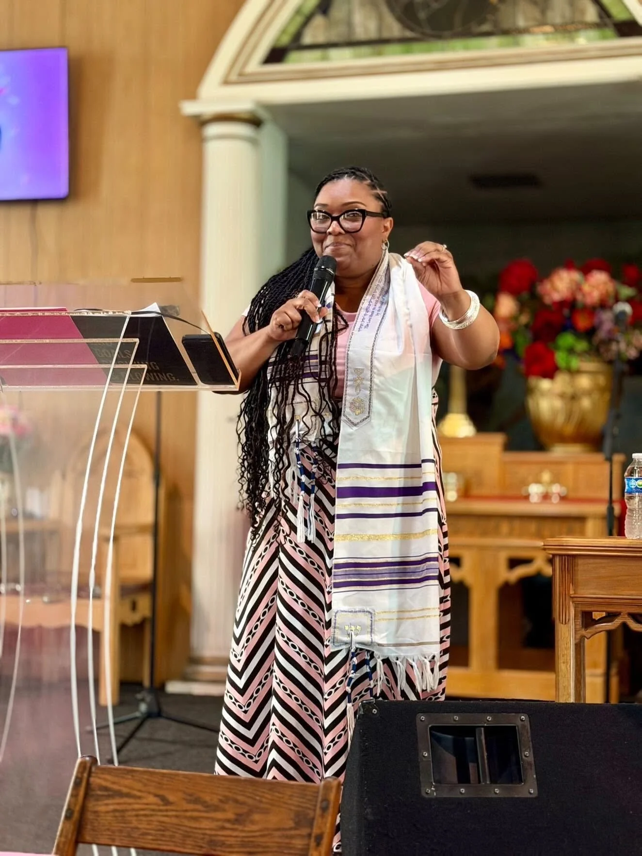 Woman with glasses, braided hair, and jewelry speaking into a microphone inside a church or community hall, wearing a colorful striped dress and a white scarf, with floral arrangements in the background.