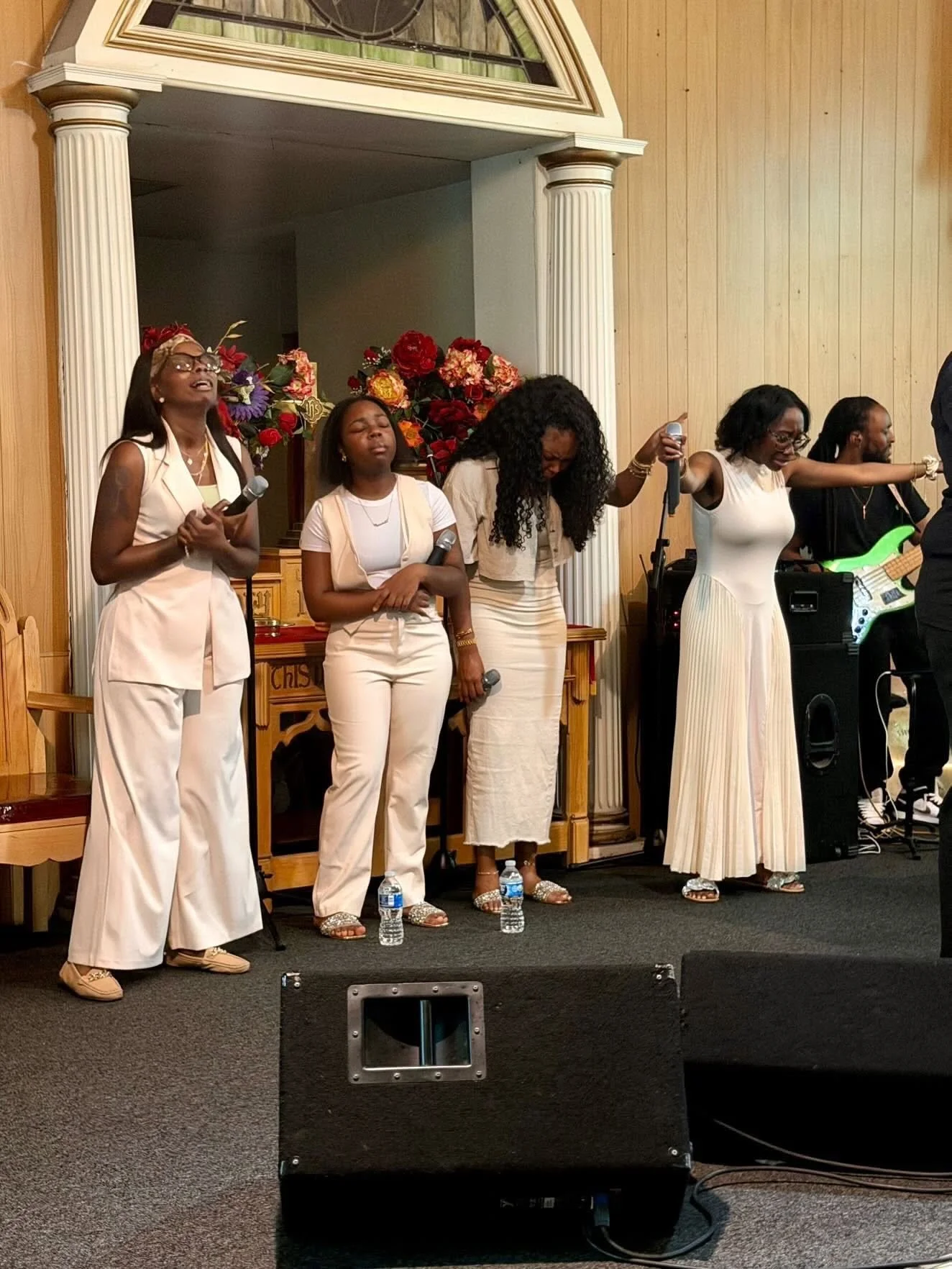 A group of five people singing or praying on a stage with a wooden background, floral arrangements, and a water bottle on the floor.