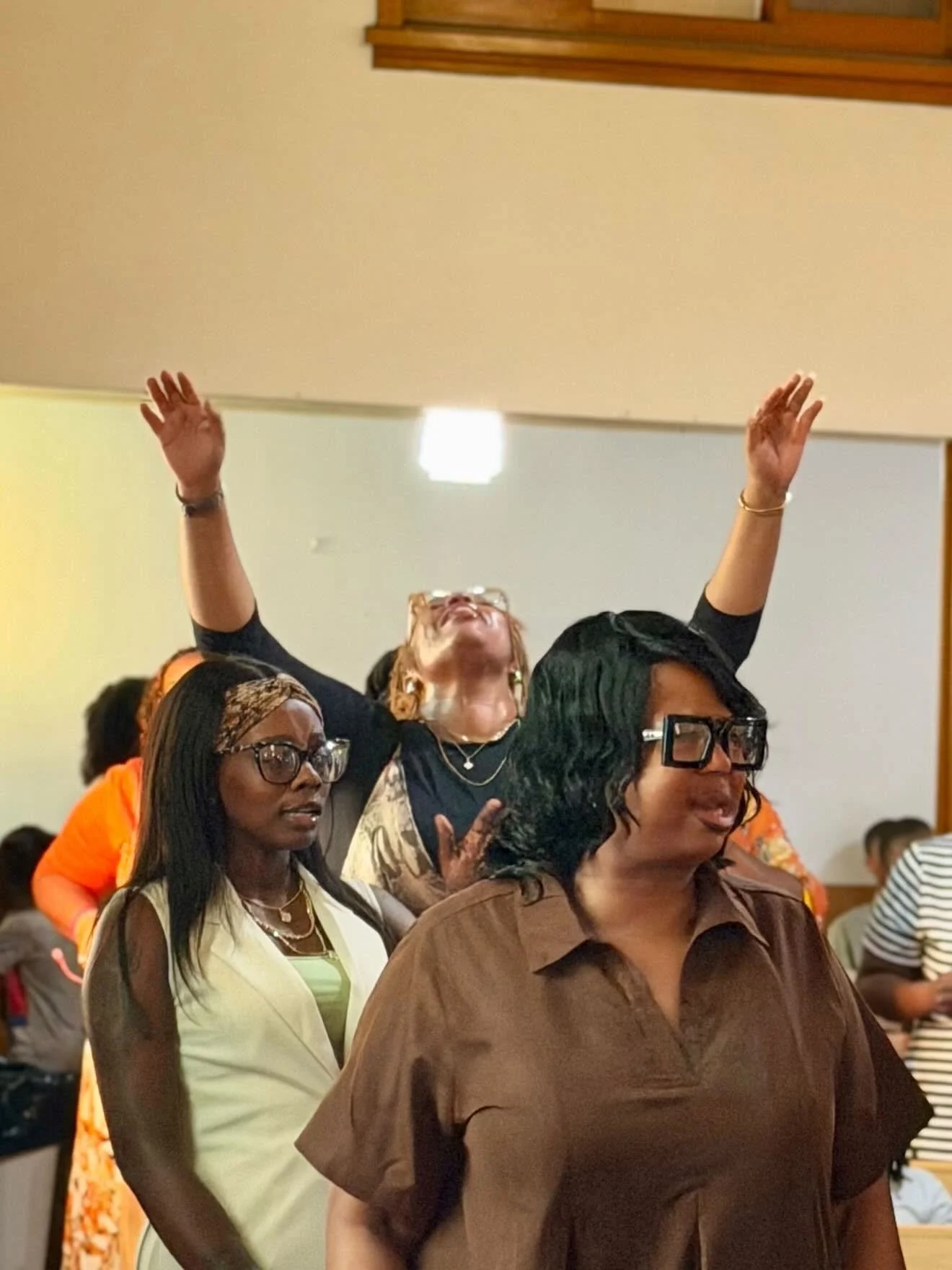 A group of women standing close together, with their eyes closed and hands raised, appearing to be engaged in a moment of prayer or worship indoors.