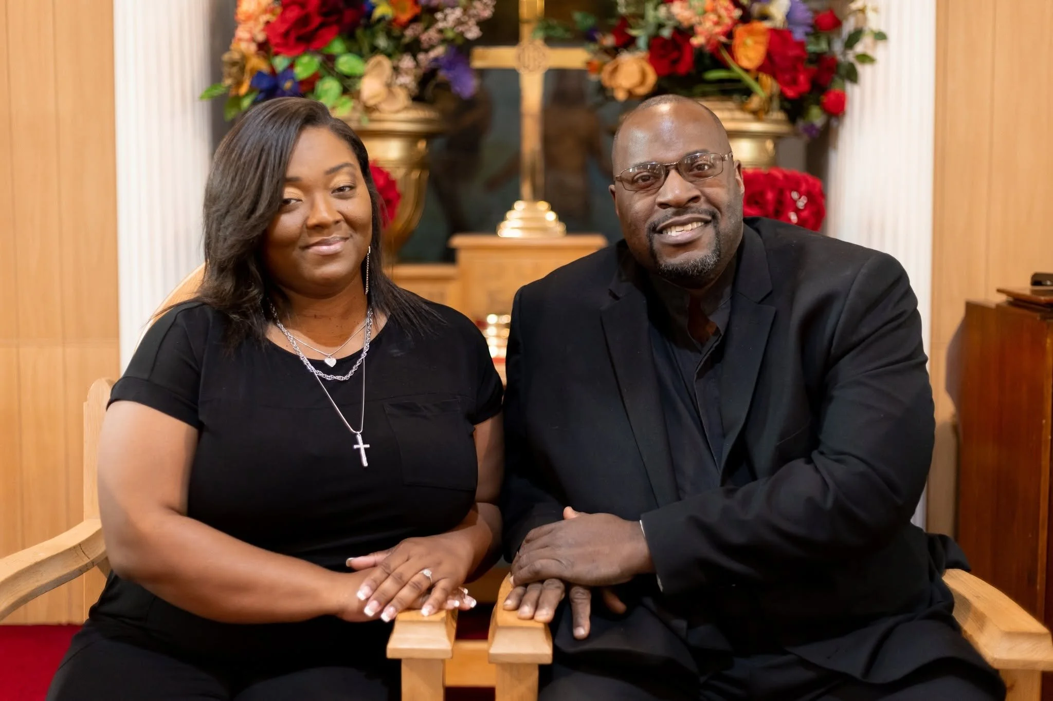 A man and a woman sitting in a church or chapel with a wooden cross and colorful flowers behind them. They are dressed in black and are smiling gently at the camera.