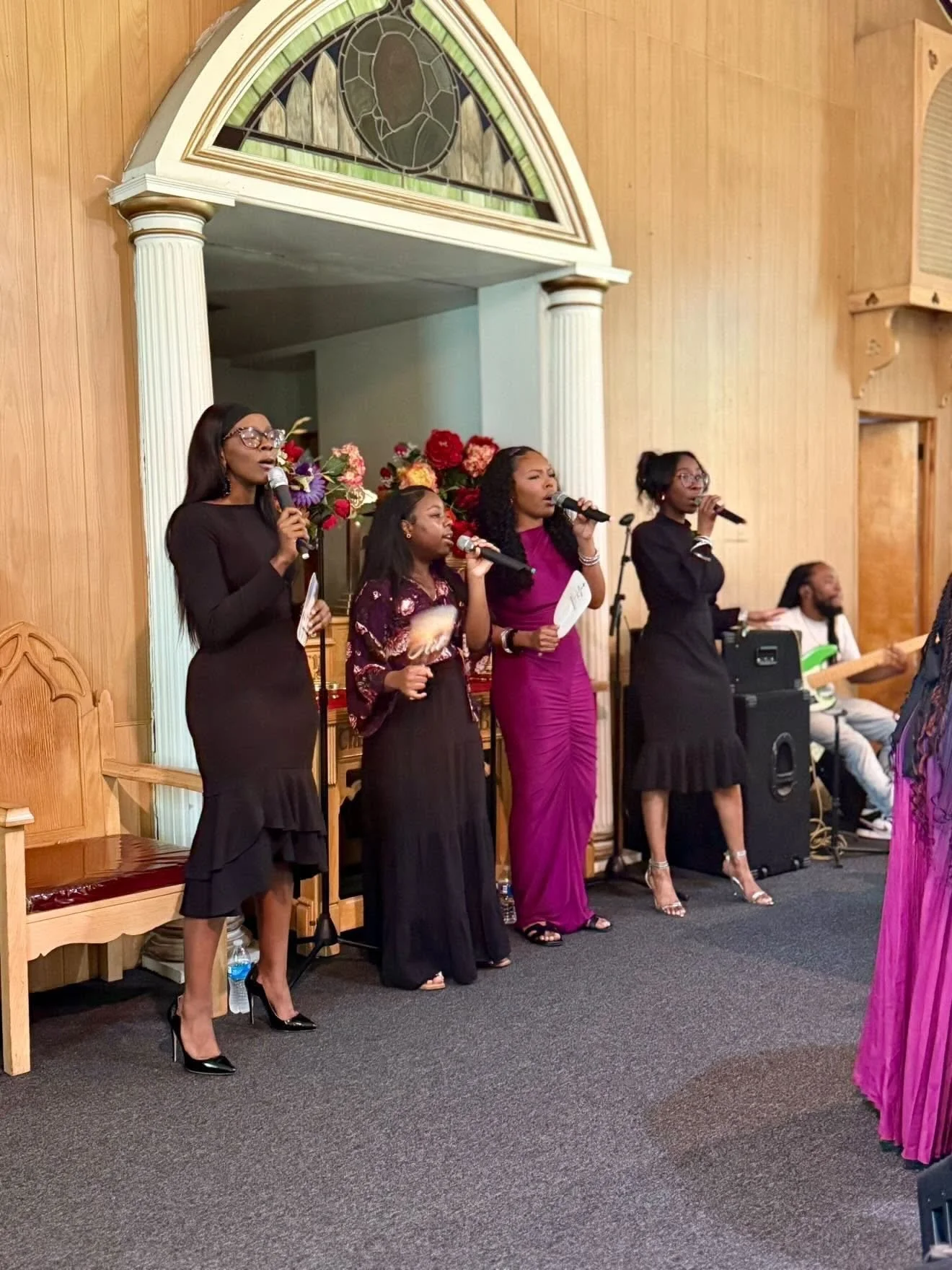 Four women singing into microphones on stage in a church setting with wooden walls and stained glass window above. Two additional musicians are in the background playing an electronic keyboard and bass guitar.