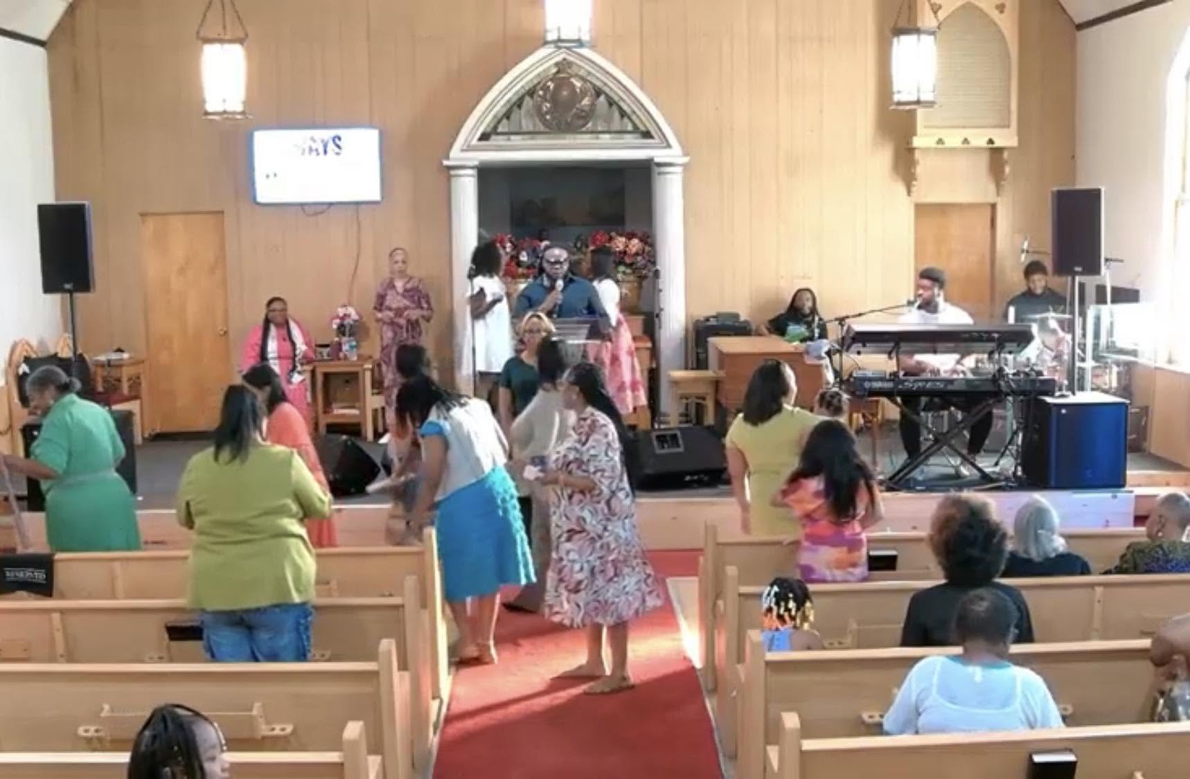 People gather in a church sanctuary with wooden walls, many of them standing and conversing. A man with a microphone stands at a lectern by an altar decorated with flowers. Several singers and musicians are positioned on the stage, with some seated at musical equipment. Windows and hanging light fixtures are visible.