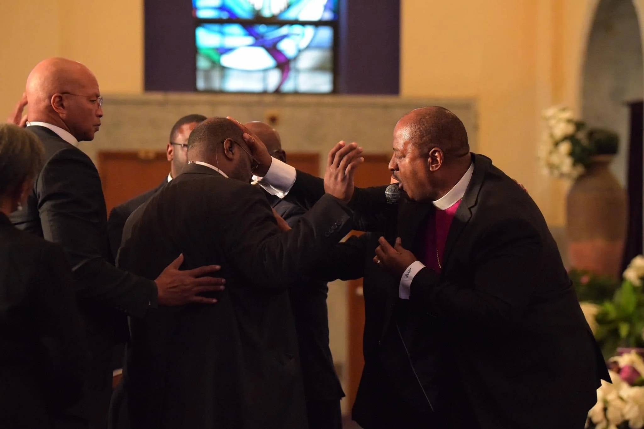 A group of men dressed in formal attire, including suits and clergy robes, are gathered inside a church. One man appears to be praying or speaking, with his hand raised and touching his forehead. The background shows a colorful stained glass window and a decorated altar.
