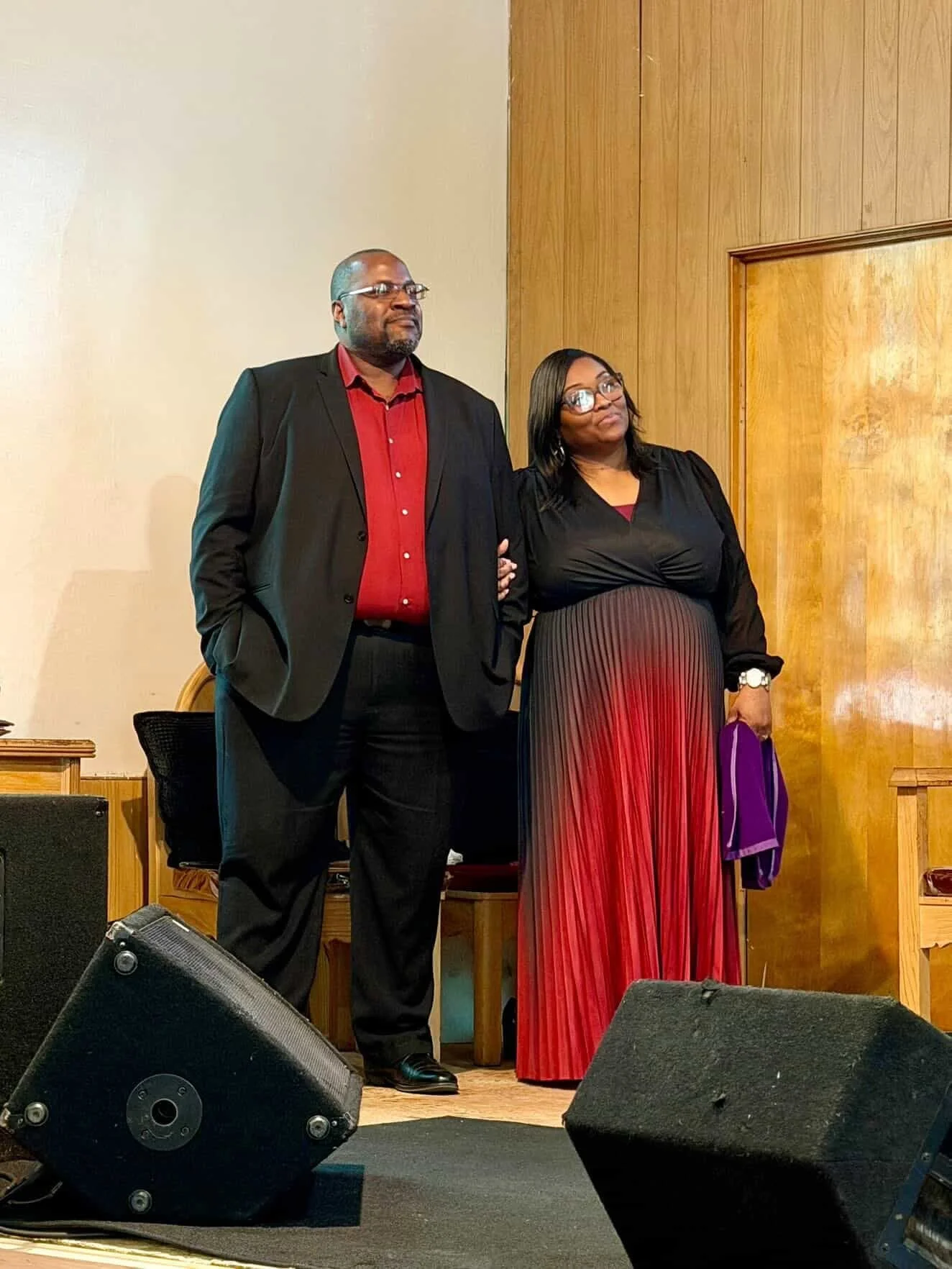 A man and a woman standing together on a stage in front of a wooden wall. The man is wearing a black suit with a red shirt, and the woman is dressed in a long, pleated red and black dress with glasses. The woman is holding a purple cloth.