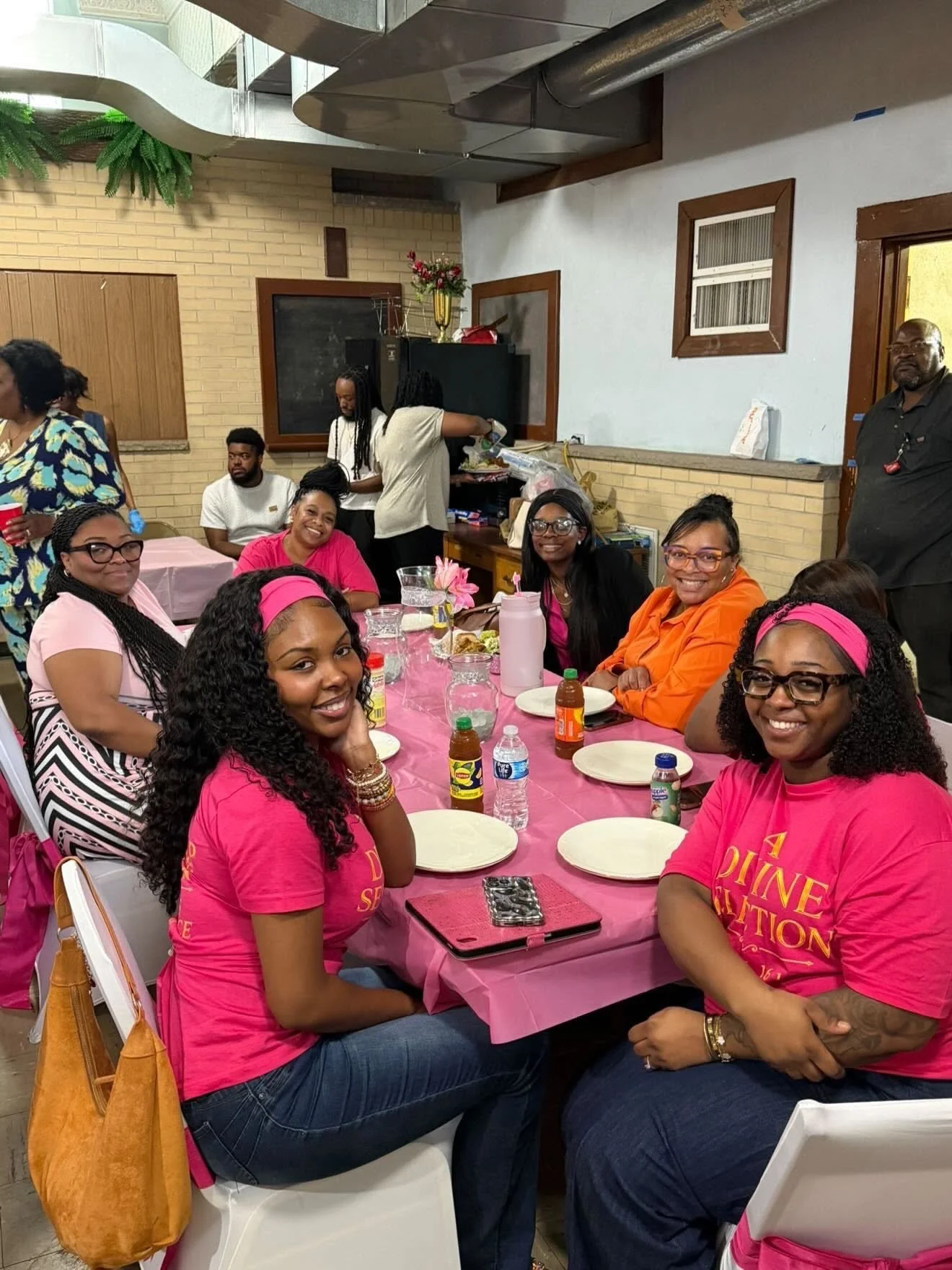 Group of women sitting around a table with pink tablecloth, smiling and looking at the camera during a celebration or gathering.