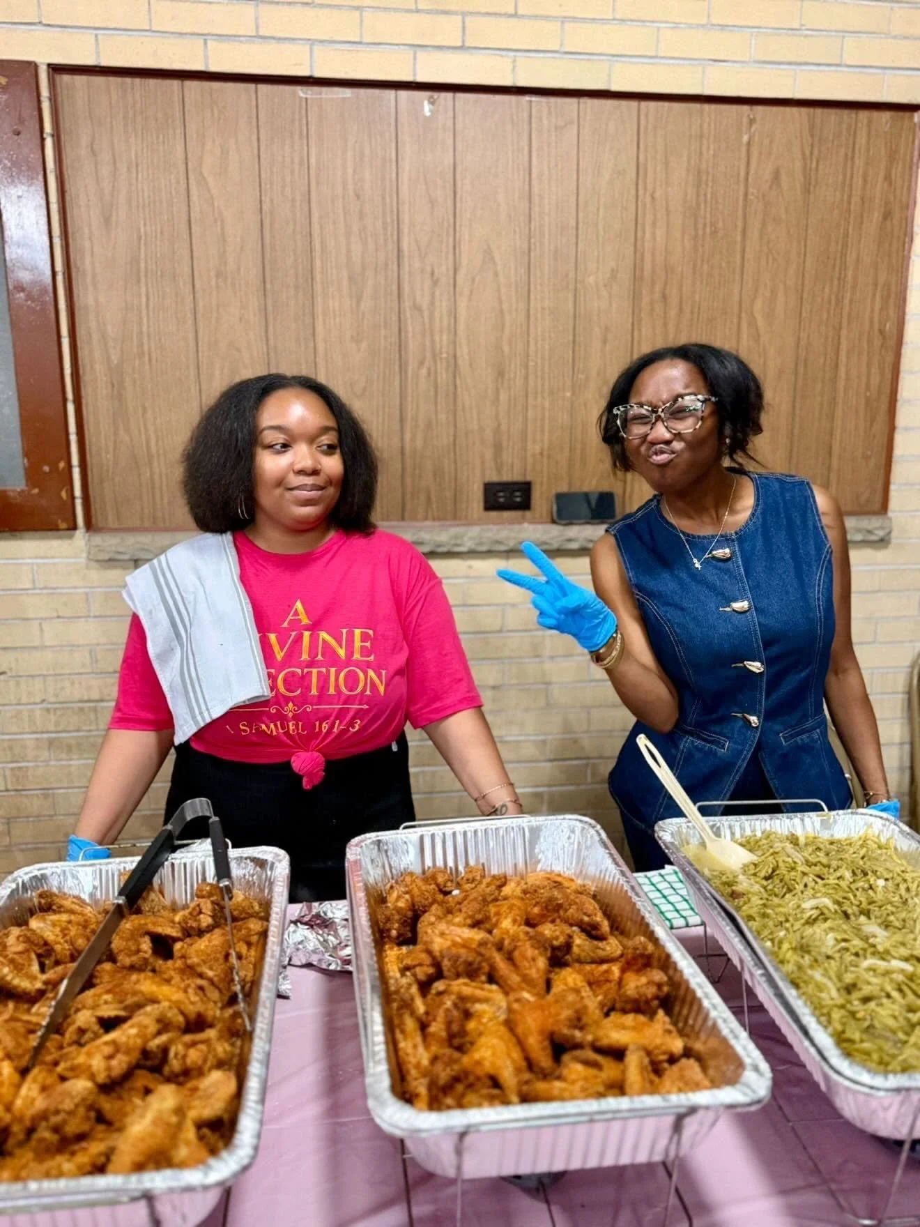 Two women standing behind a table with trays of fried chicken, noodles, and other food. One woman is wearing a red shirt with text and has a towel over her shoulder. The other woman is wearing glasses, a denim vest, and gloves, making a playful face and peace sign.