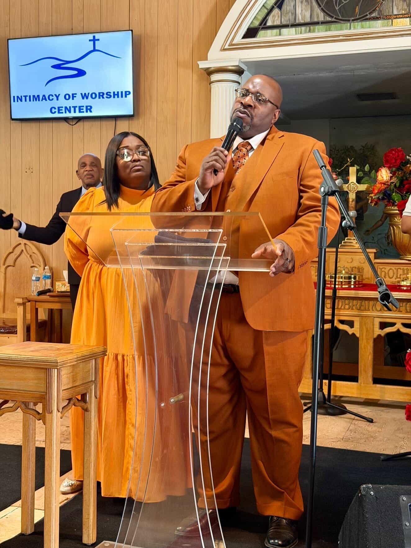 A man in a brown suit speaking into a microphone at a church, with a woman in a matching brown dress standing behind him. The church interior includes flowers, a wooden table, and a digital screen displaying 'Intimacy of Worship Center' with a cross and hill graphic.
