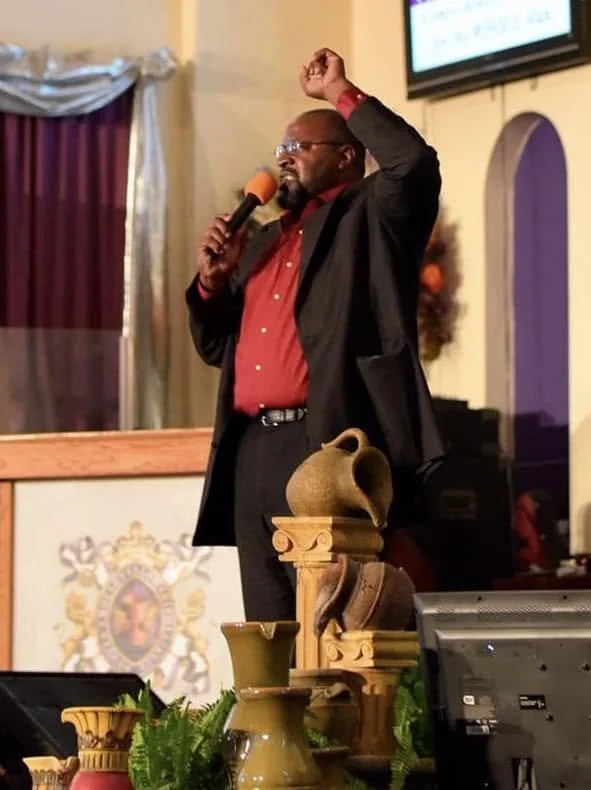 A man in a dark suit and red shirt is speaking into a microphone with a yellow cover, raising his right hand in a gesture of emphasis, standing behind a display of various pottery vases on a table. The setting appears to be a church or auditorium with purple curtains and a decorative setting.