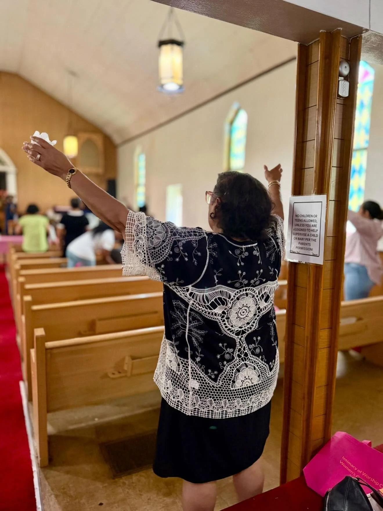 A woman in a black and white embroidered dress stands with her arms raised inside a church. There are wooden pews and stained glass windows in the background. A sign on a wooden post restricts children without supervision.