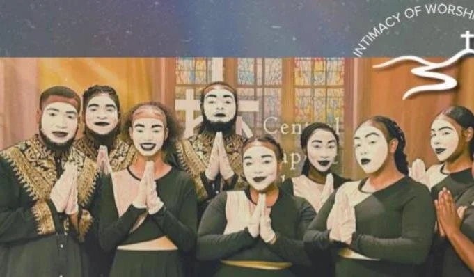 Group of people with painted faces posing inside a church or religious space, some with hands in prayer position, with stained glass windows in the background.