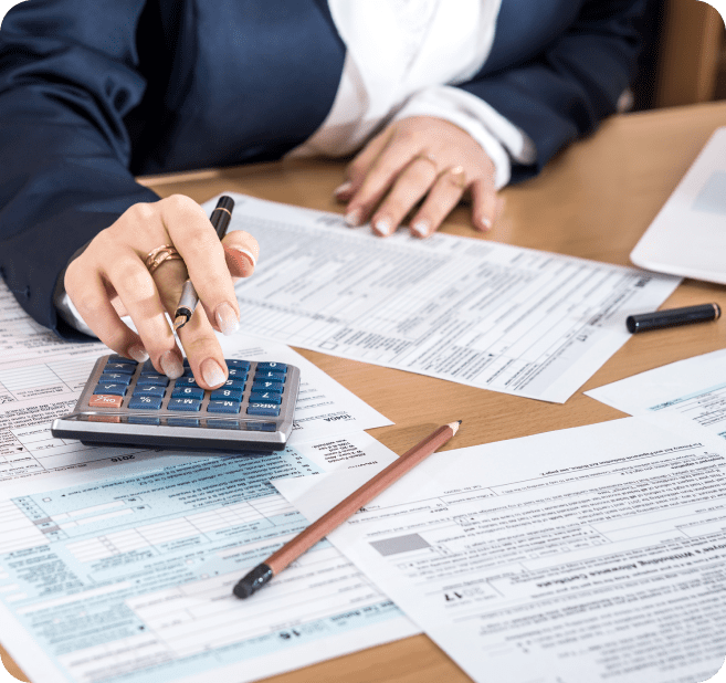 Person in business attire using a calculator and pen on a cluttered desk with documents and a laptop.