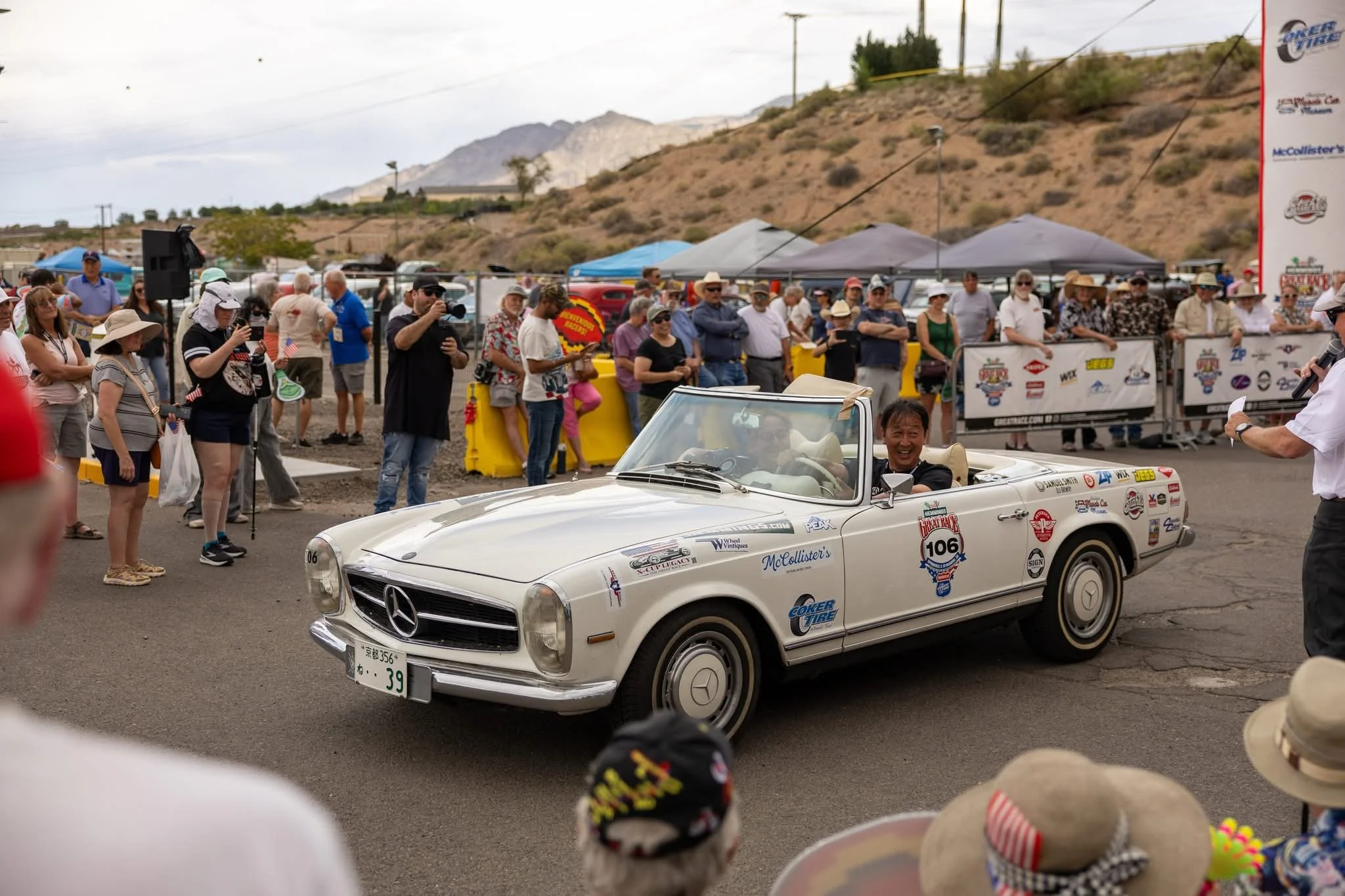 A vintage white Mercedes-Benz convertible with event stickers and the number 106 on the door, driving past a crowd of people at an outdoor event, with tents and mountainous landscape in the background.