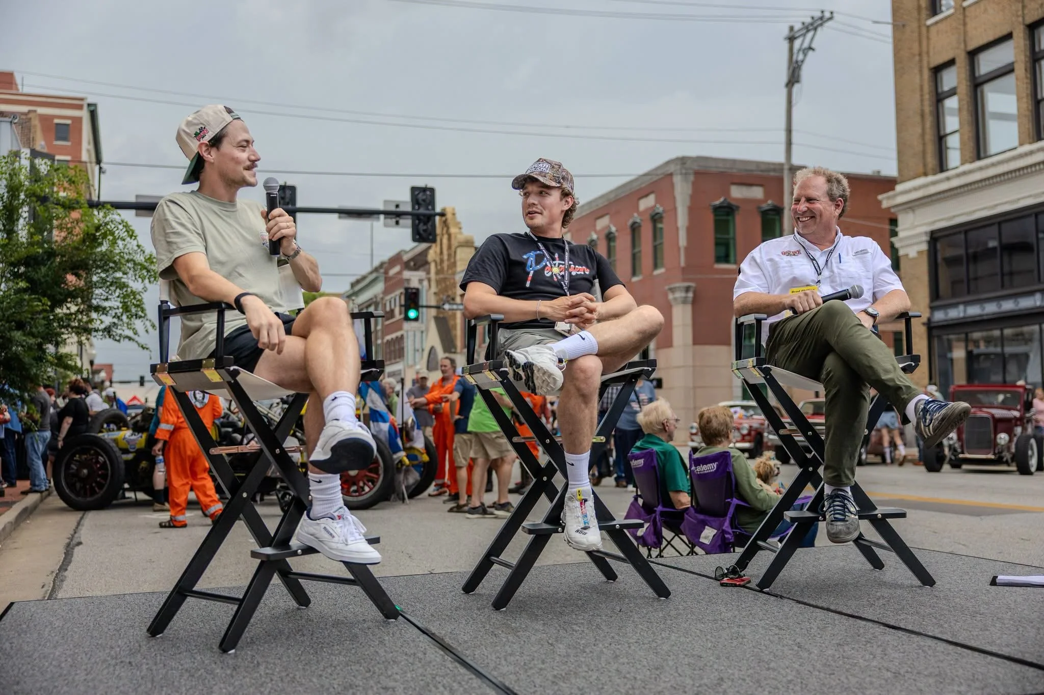 Three men sitting on chairs on a street stage during an outdoor event, engaging in a panel discussion. The man on the left holds a microphone, wearing a beige T-shirt and cap. The man in the middle wears a black T-shirt and cap, and the man on the right wears a white shirt and khaki pants, smiling. There are people and buildings in the background, with a crowd gathered around.