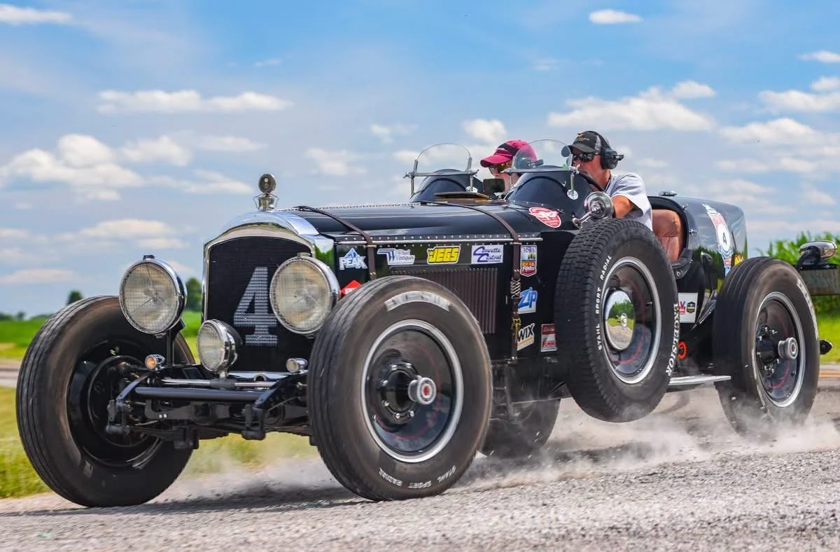 A vintage black race car with the number 4 on the front, driving on a gravel road with two people wearing headphones and caps inside.