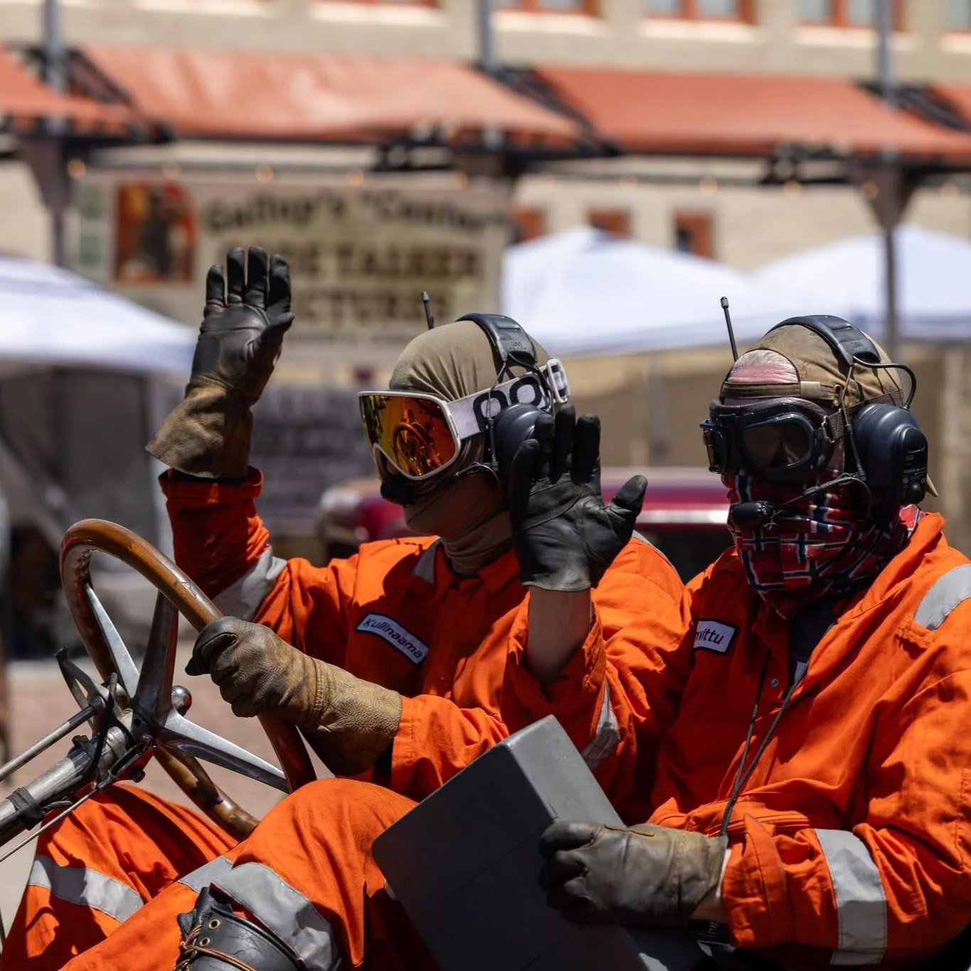 Two individuals in orange uniforms and protective gear, including helmets with headsets and goggles, are riding a vehicle. One person is waving. They are in an outdoor market or street setting.