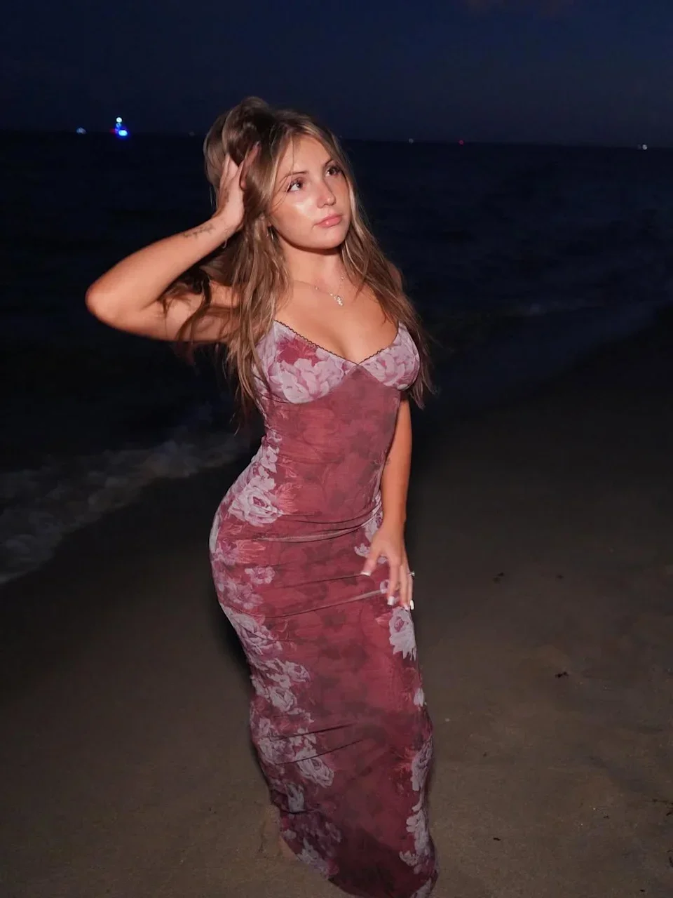 A woman in a long floral dress standing on a beach at night, with the ocean and dark sky in the background.