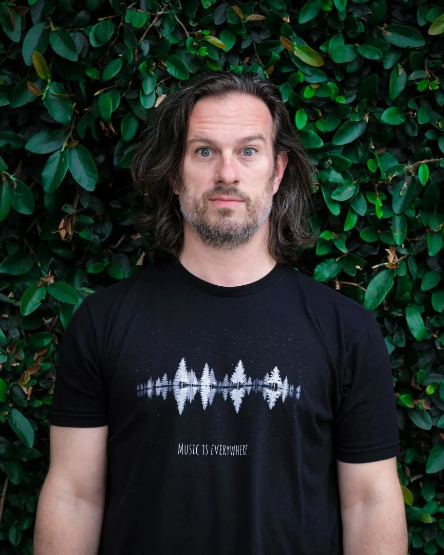 A man with long dark hair and a beard standing in front of green leafy bushes, wearing a black T-shirt with a tree reflection graphic and the words "Music is Everywhere" on it.