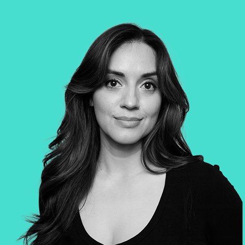 A woman with long wavy hair taking a selfie, wearing a black top, against a turquoise background.