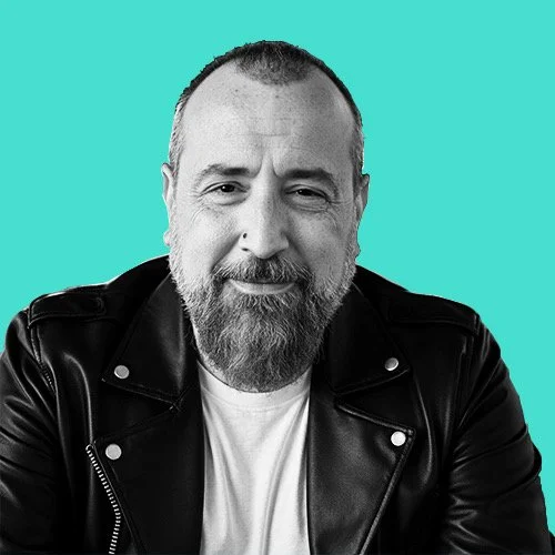 A middle-aged man with a beard and short hair, wearing a black leather jacket and a white t-shirt, smiling against a solid teal background.