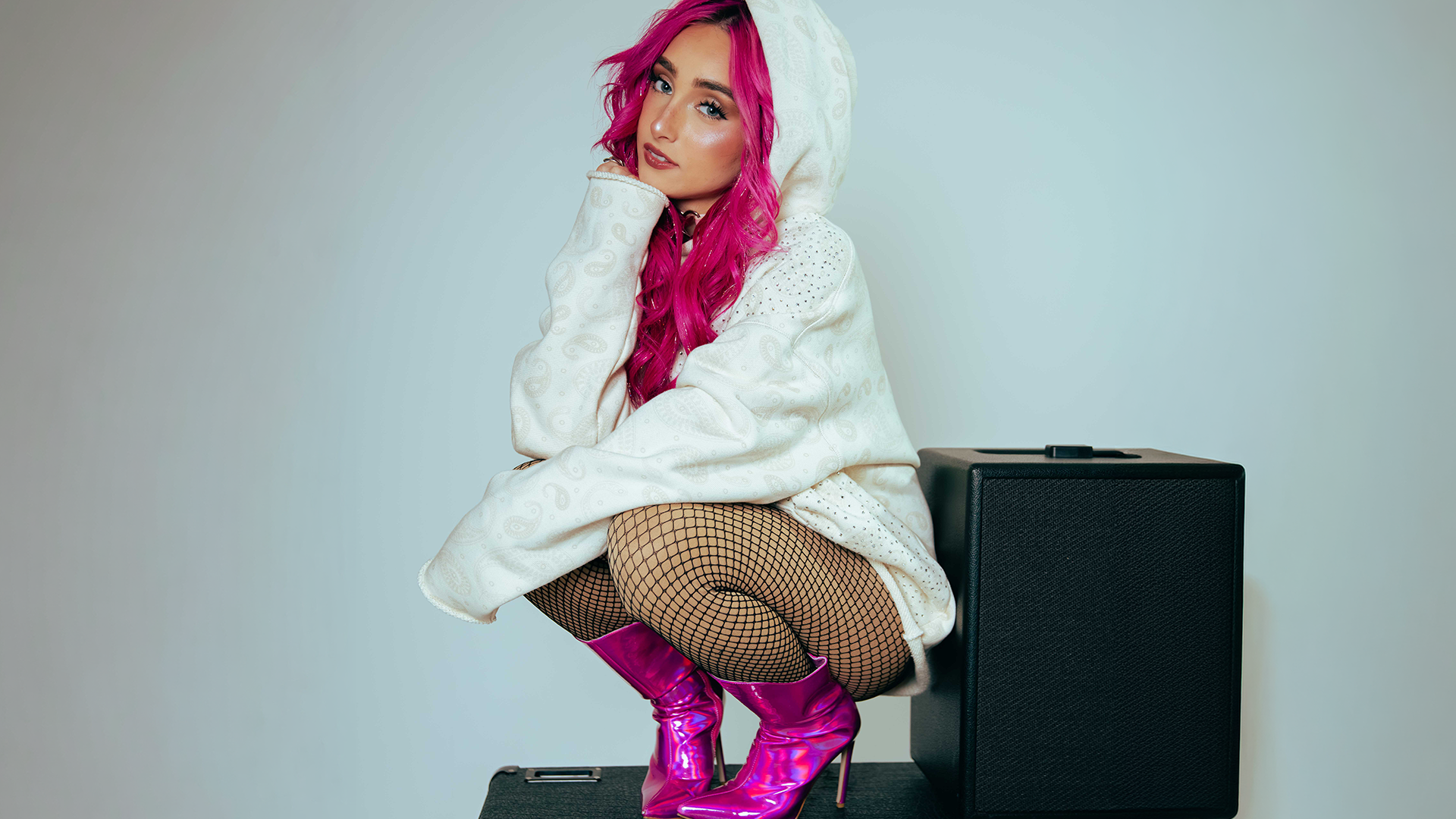 Young woman with bright pink hair sitting on a speaker against a plain white wall, wearing a baseball cap, oversized hoodie, fishnet stockings, and shiny pink boots.