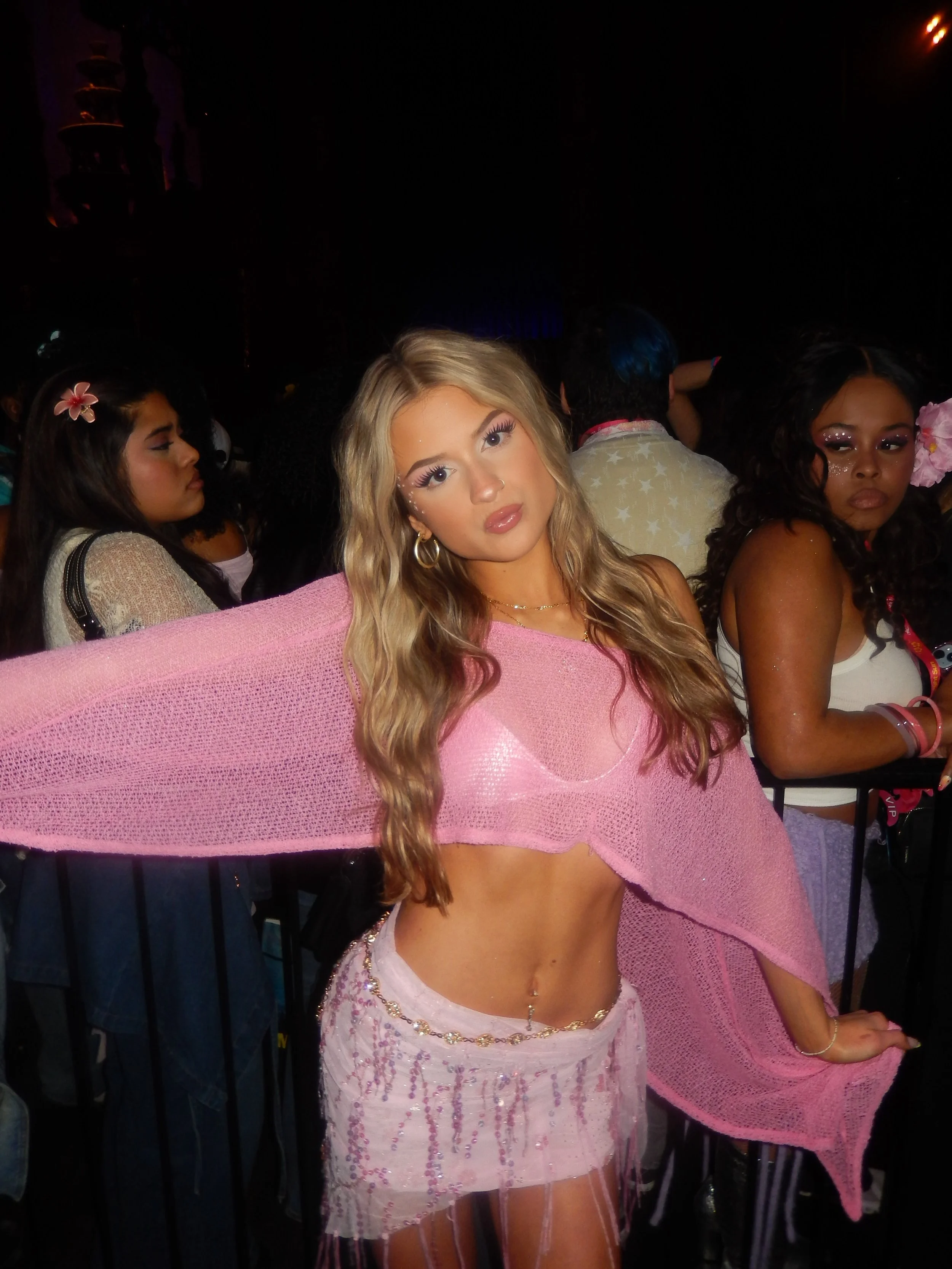A woman with long blonde hair and makeup, wearing a sheer pink top and white skirt with pink accents and fishnet stockings, is at a party. She is in front of a group of women, some with flowers in their hair, in a dimly lit environment.