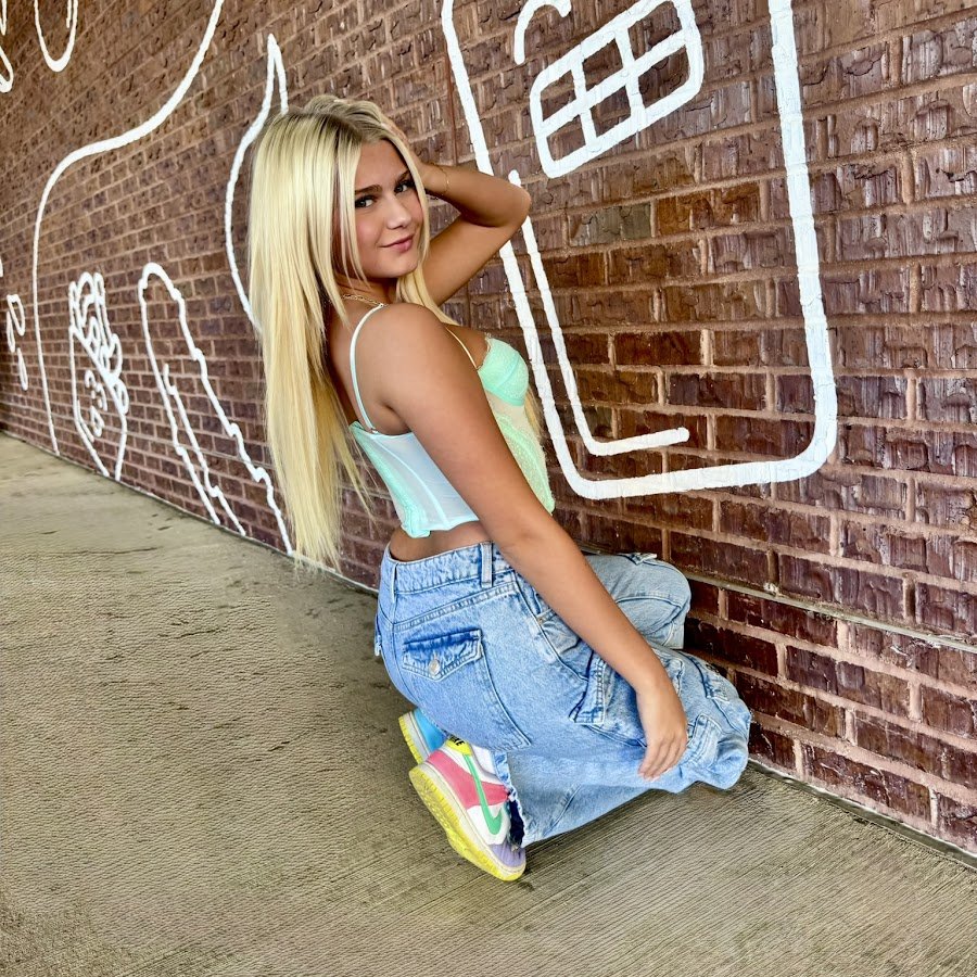 Young woman with long blonde hair wearing a mint green crop top, baggy jeans, and colorful sneakers, sitting on her knees against a brick wall with white chalk drawings of a house and other shapes.