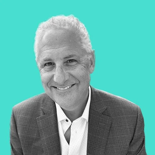 Black and white portrait of a middle-aged man with gray curly hair, smiling, wearing a suit jacket and a white shirt, with a turquoise background.