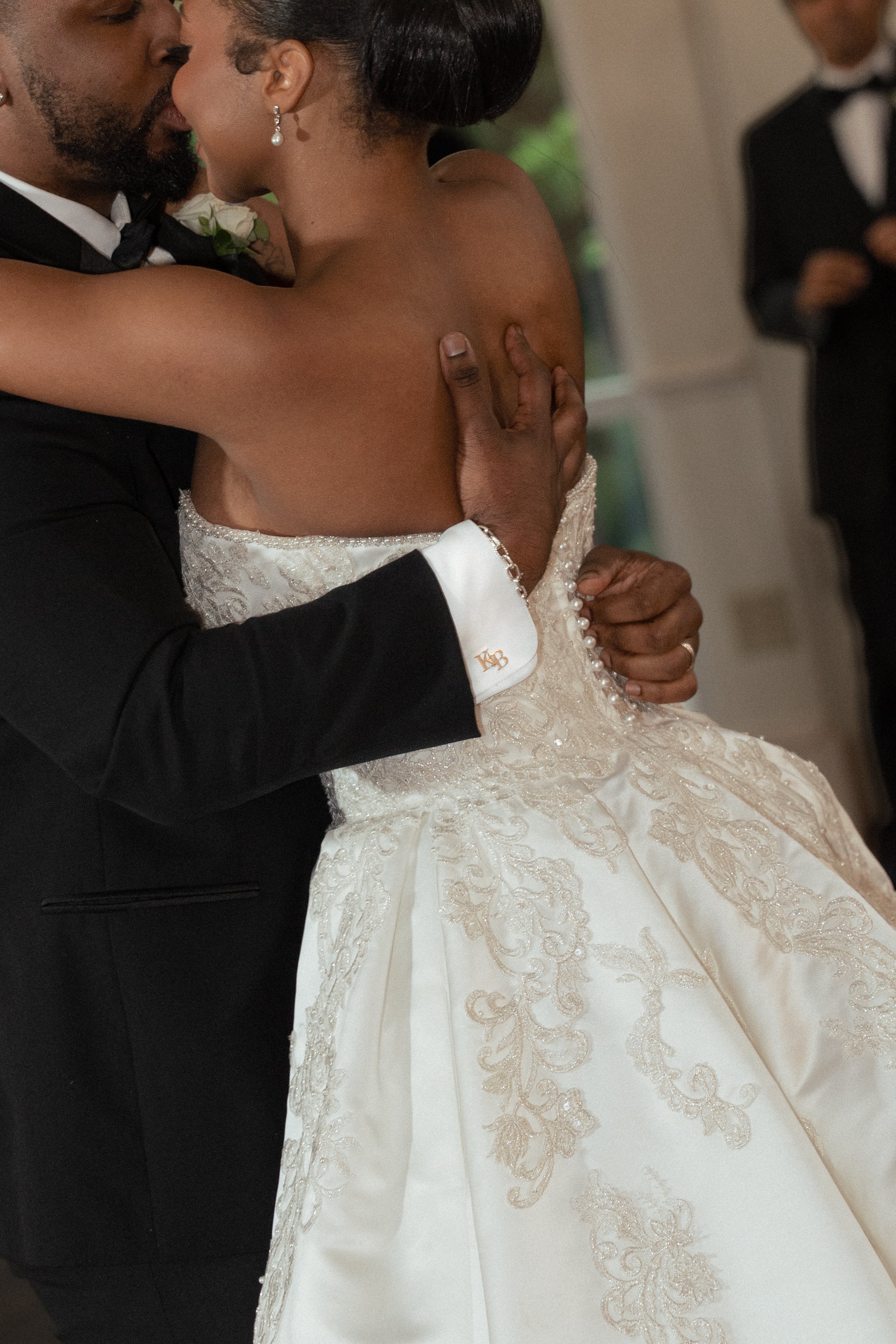 A close-up of a wedding kiss between a man in a black tuxedo and a woman in a strapless white wedding dress with intricate embroidery, as a guest in formal attire watches in the background. Shot in Ashton Gardens in Sugarhill, Georgia